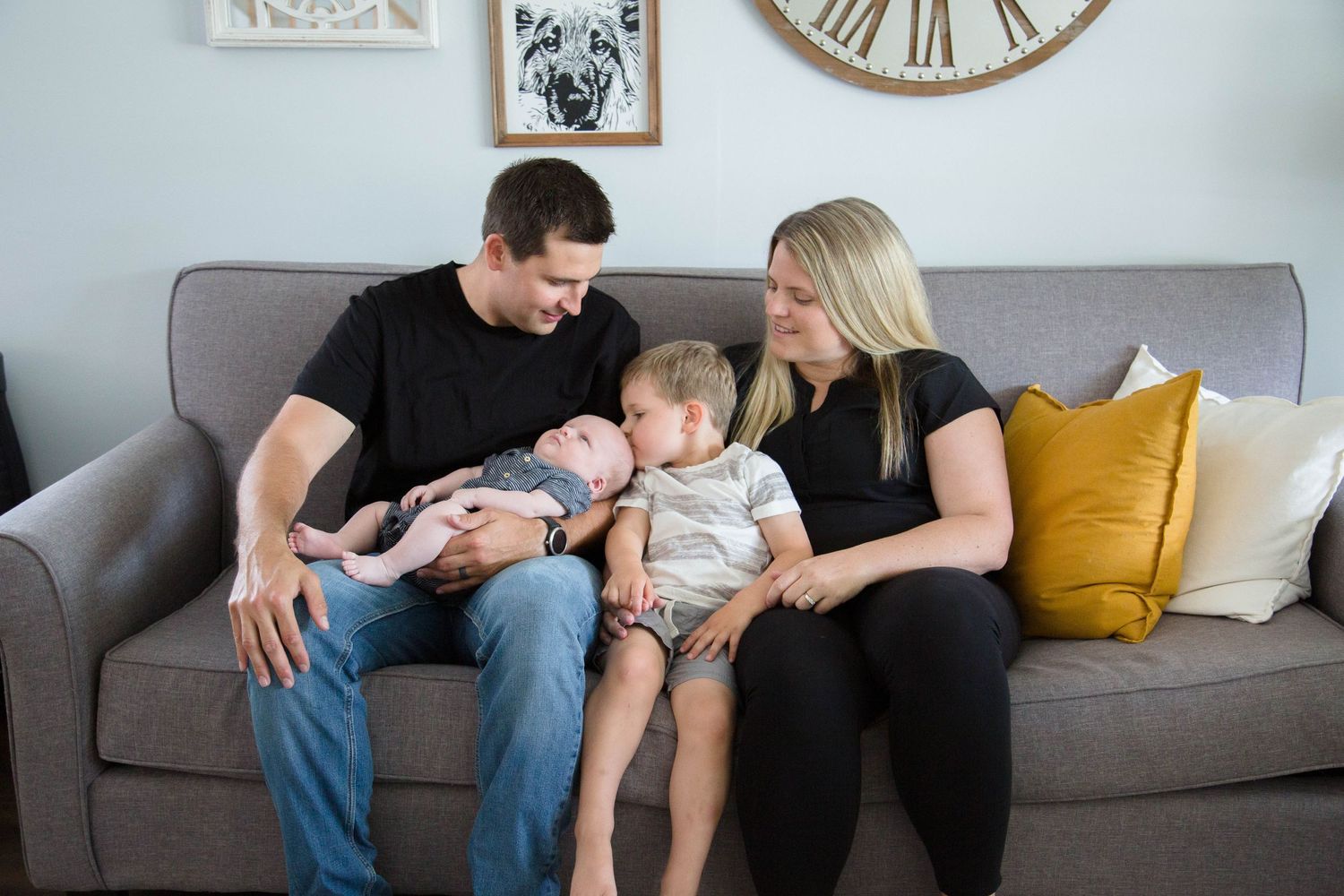 Parents with toddler and newborn baby bonding together on gray sofa in cozy living room.