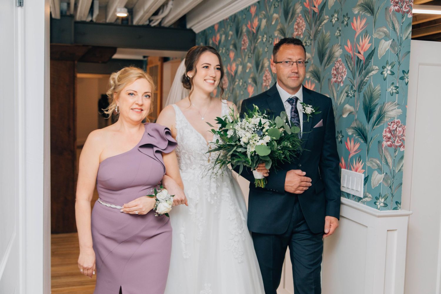Bride with parents walking down aisle in floral-wallpapered wedding venue.