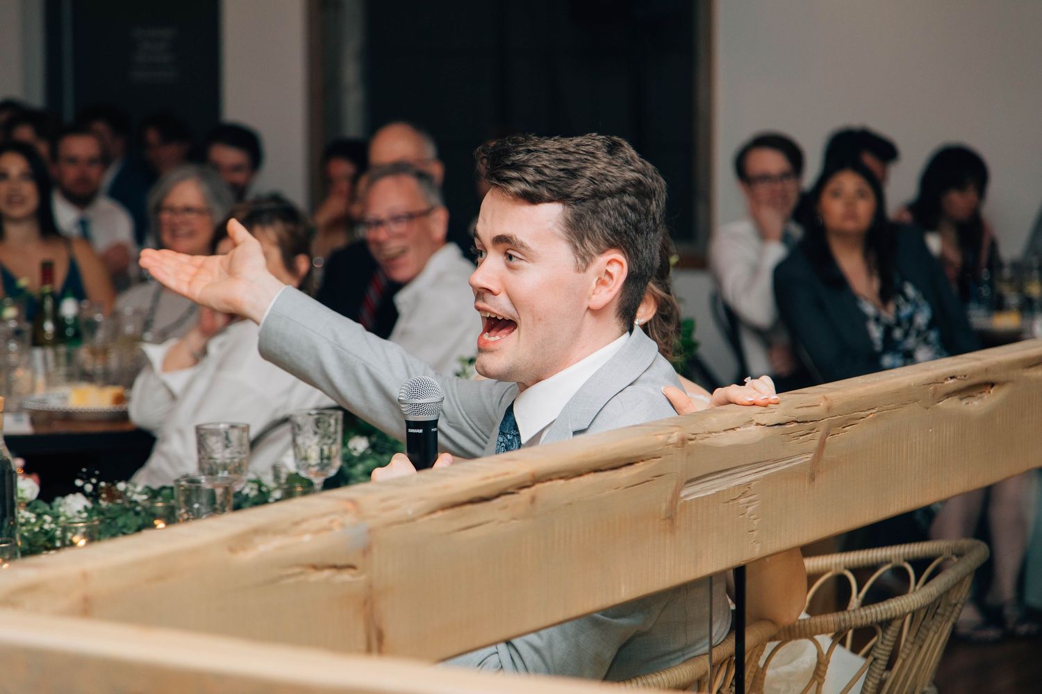 Excited groomsman in gray suit giving an animated wedding toast speech to reception guests.