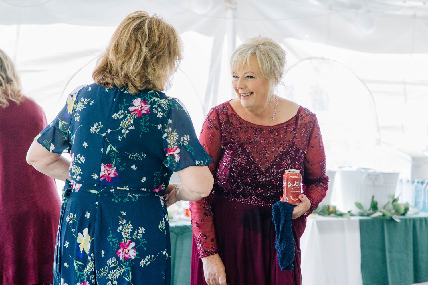 Two women in festive dresses chat and laugh together under a white tent at a wedding reception.