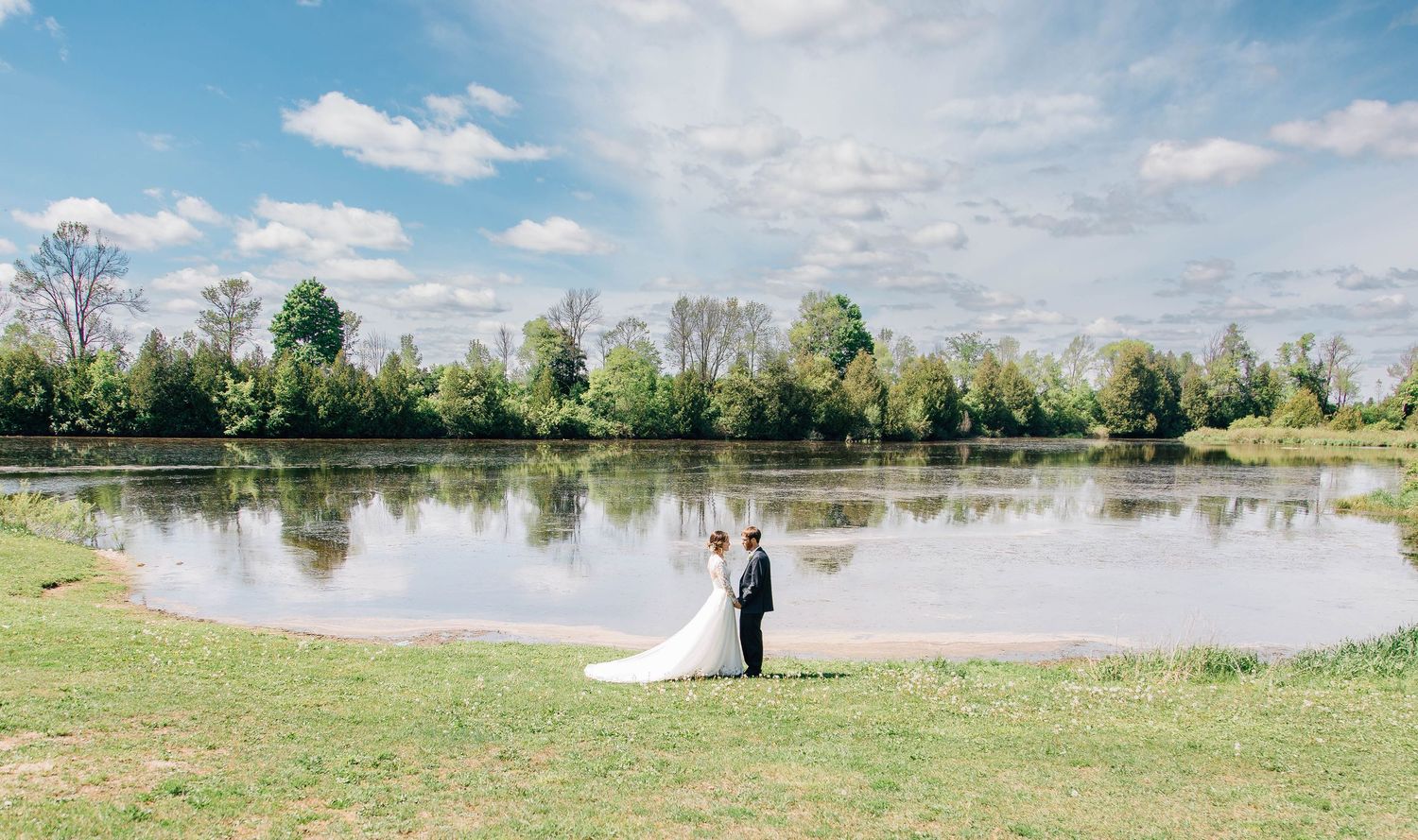 Wedding couple standing together by a peaceful lake surrounded by lush green trees.