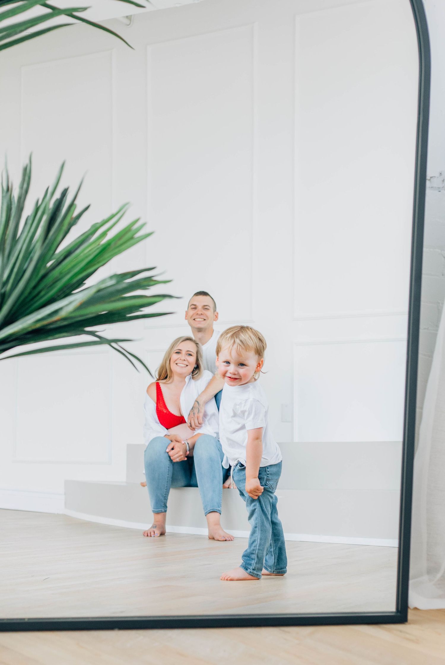 Creative mirror reflection maternity photo of family with toddler walking in bright white studio space.