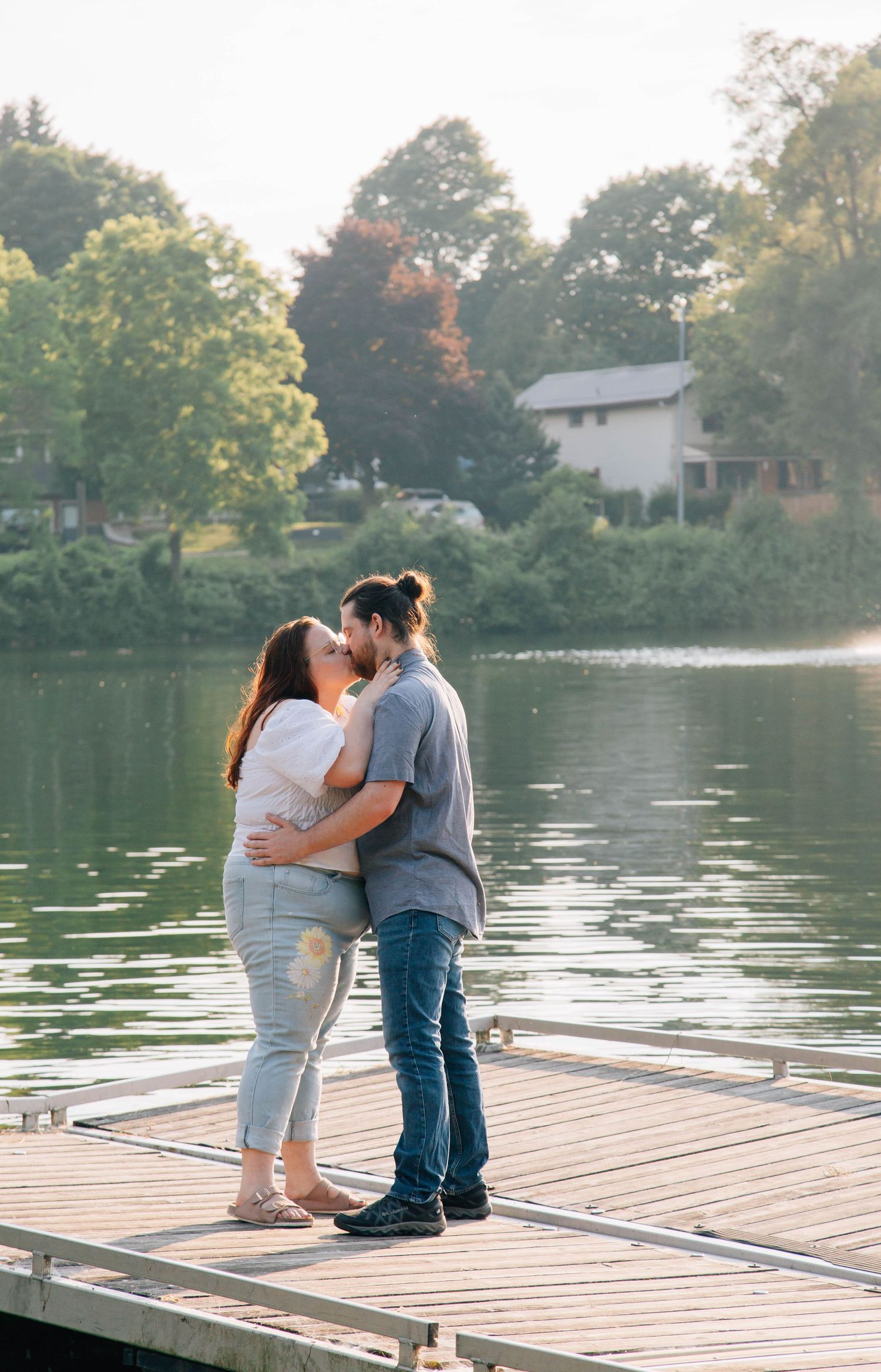Couple sharing a romantic kiss on a wooden dock with a peaceful lake and trees behind them.