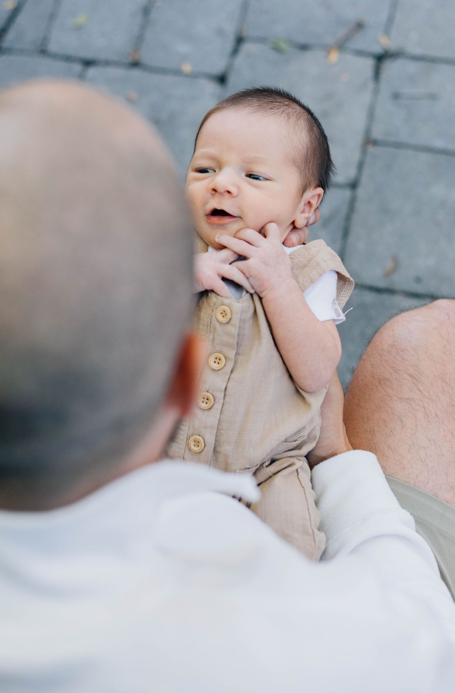 Bald father holding alert newborn baby in tan button romper on cobblestone surface, baby looking up curiously.