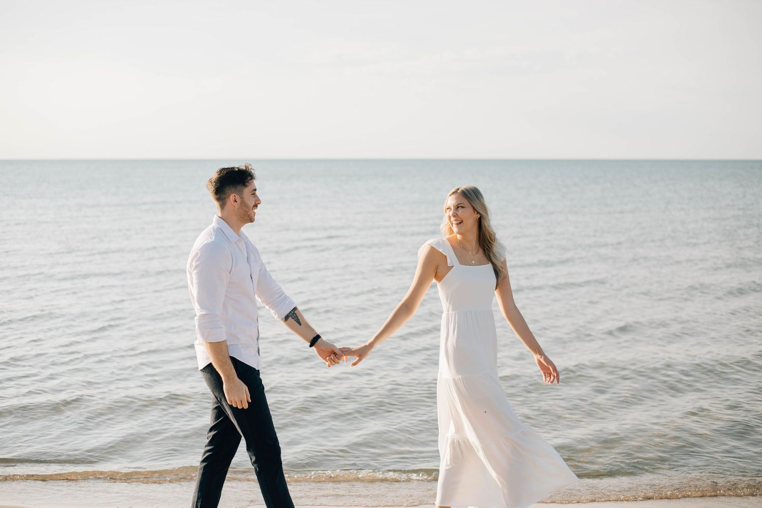Couple holds hands walking along serene ocean shoreline during romantic beach engagement photo session.