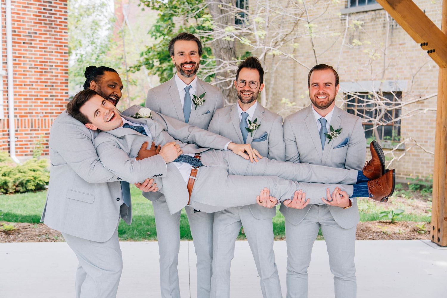 Groomsmen in gray suits playfully carry groom horizontally outdoors at wedding venue while laughing.