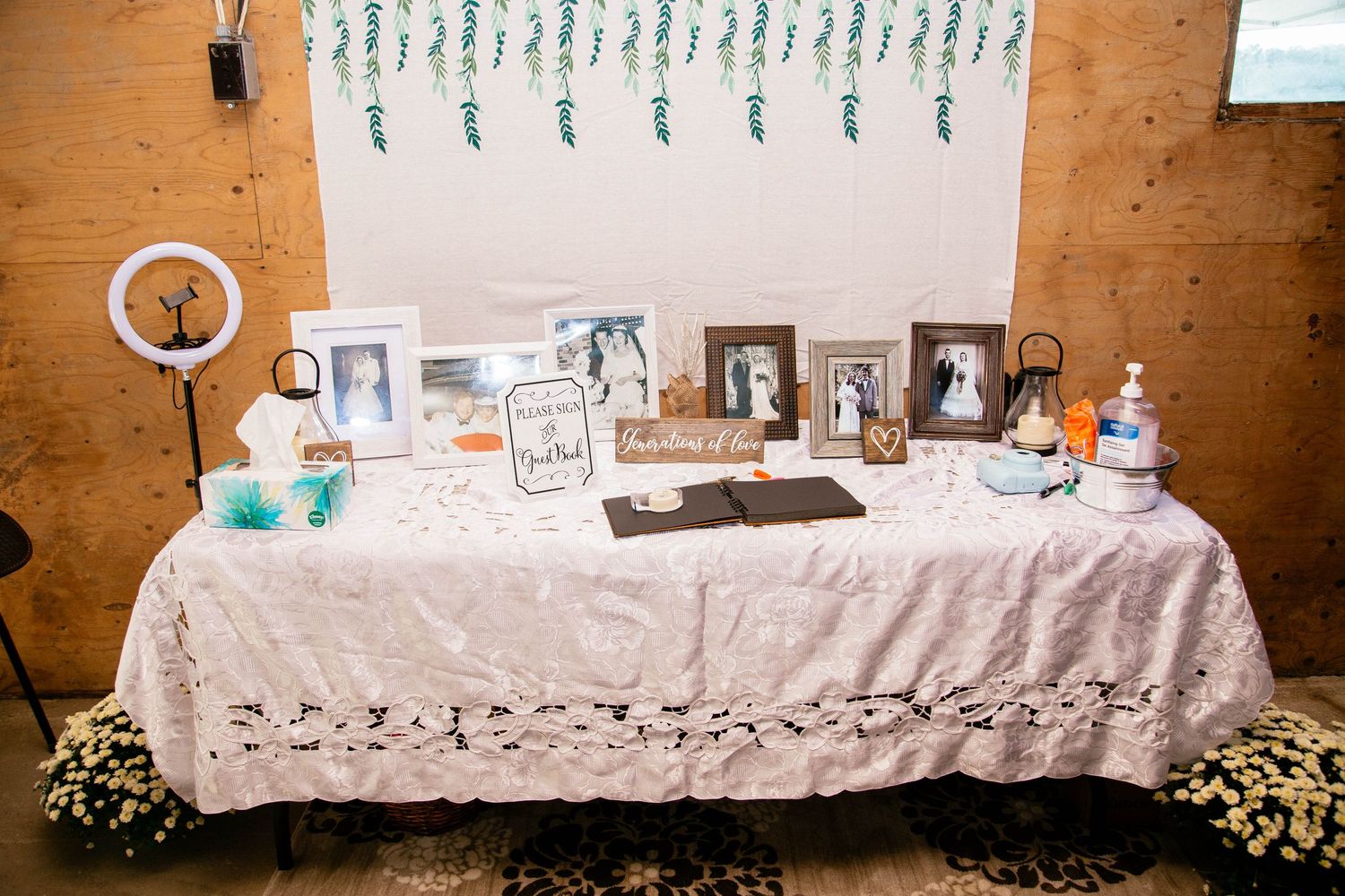 Wedding guest book table with lace tablecloth, framed photos, ring light, and floral backdrop decorations.