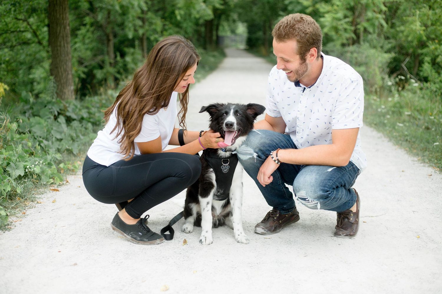 Smiling couple crouching down with their black and white Border Collie dog on a scenic gravel trail.