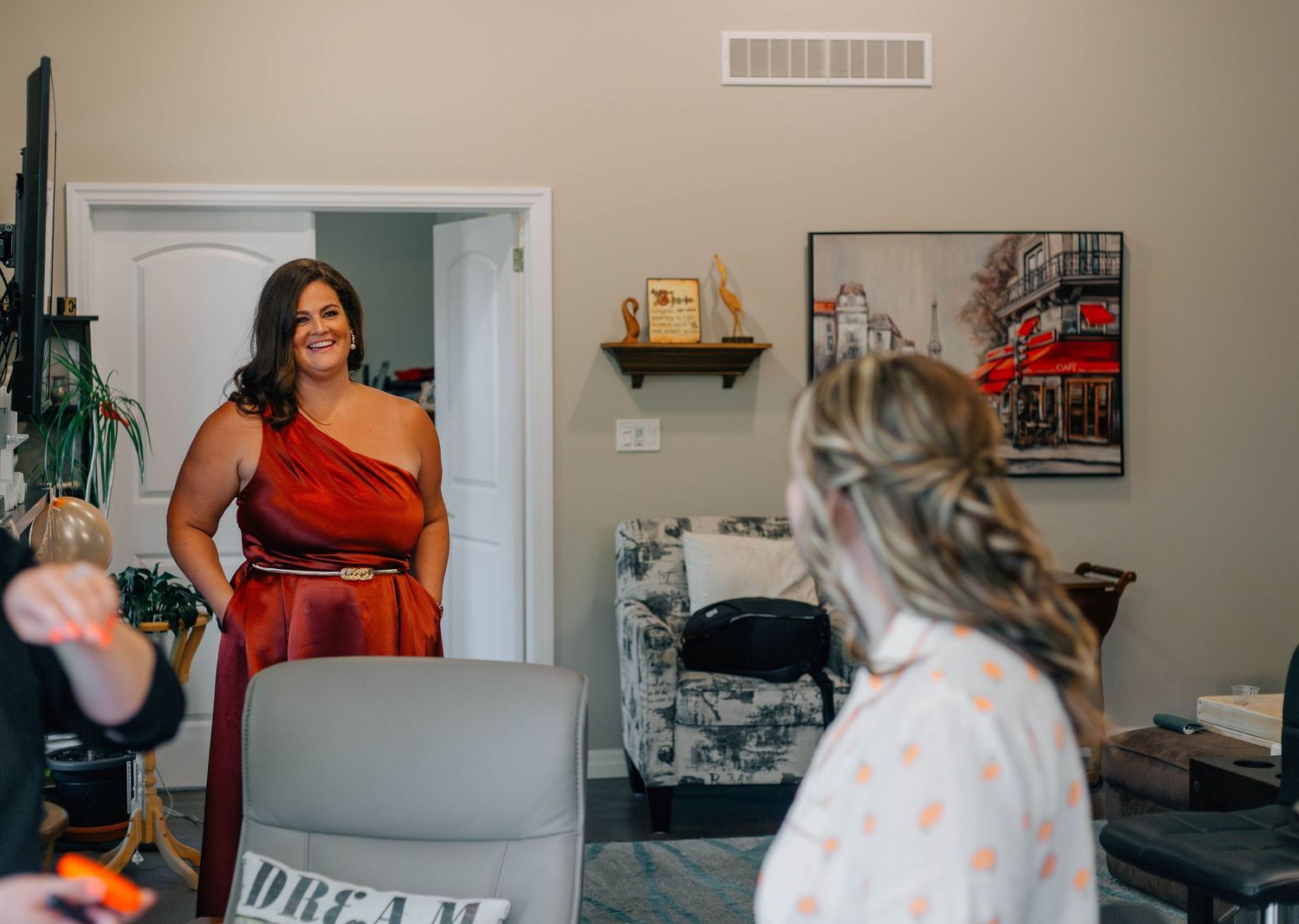 Bridesmaid in a red one-shoulder gown smiles warmly at the bride getting ready in a cozy bedroom.