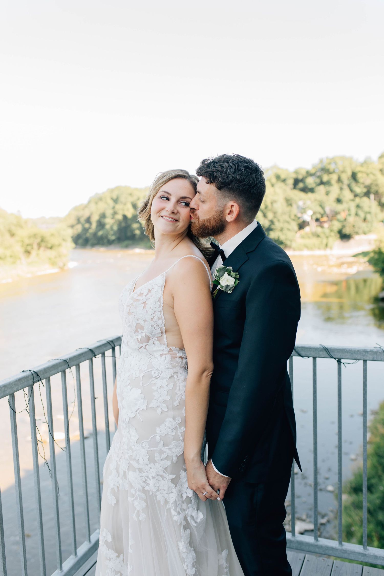 Groom in black tuxedo kisses bride in floral lace gown on bridge overlooking scenic river in Paris Ontario.