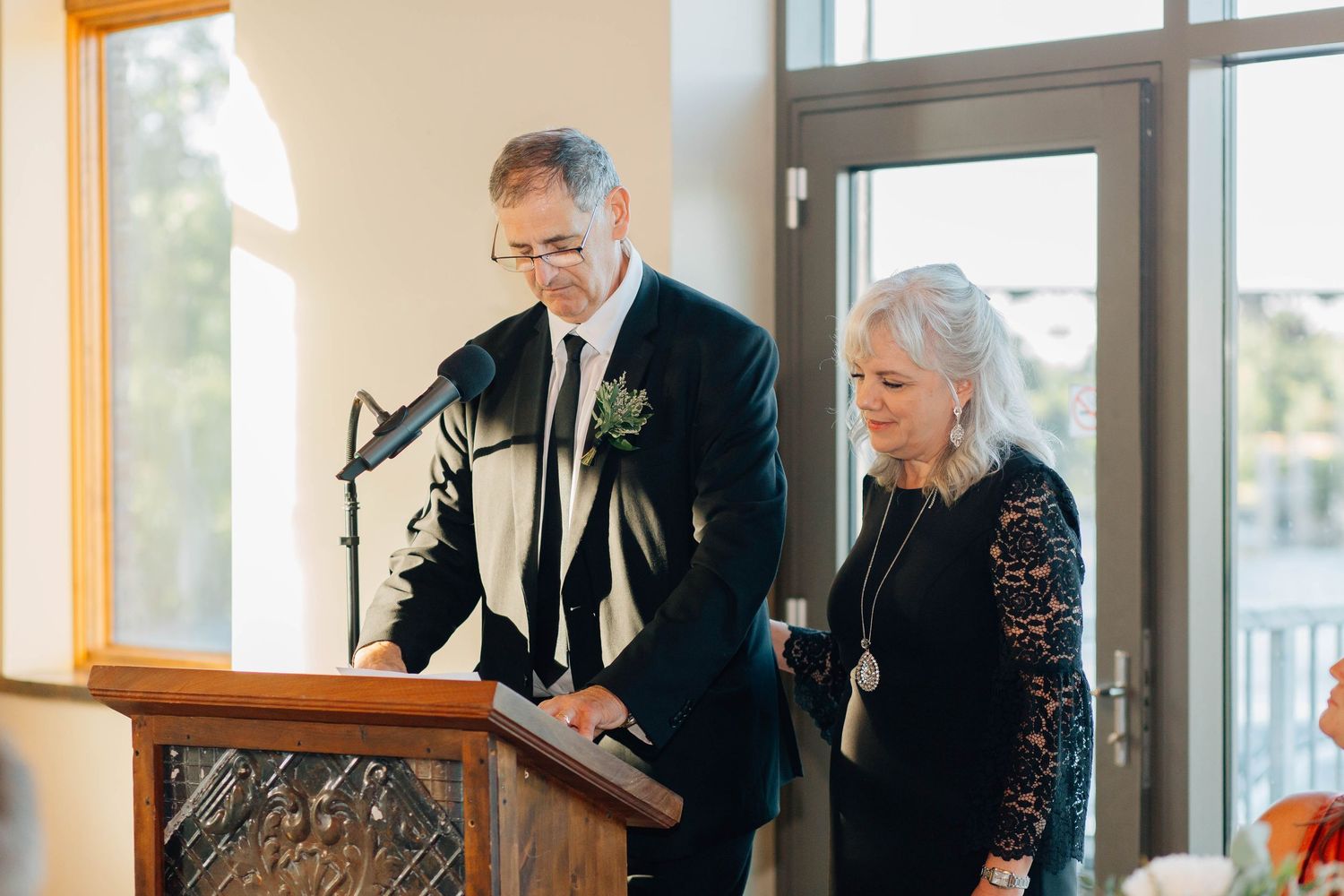 Parents of bride in formal attire reads a heartfelt speech together at a wooden podium during wedding reception.