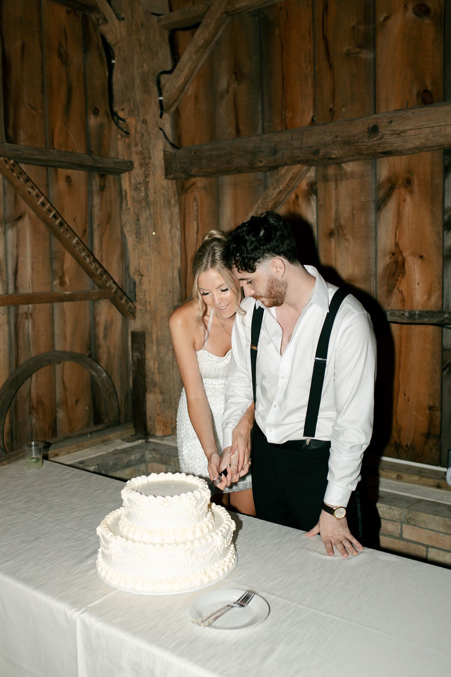 Bride and groom cutting white tiered wedding cake in rustic barn venue, woman in white dress, man in suspenders.