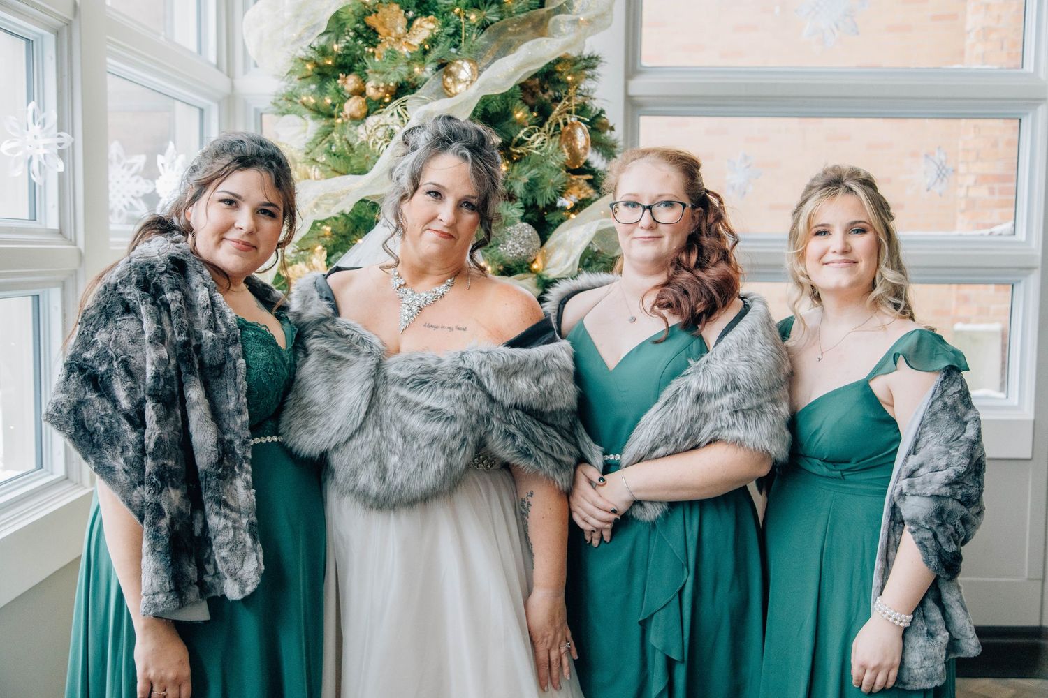 Bride with three bridesmaids in emerald green gowns and gray fur wraps posing by Christmas tree at winter wedding.