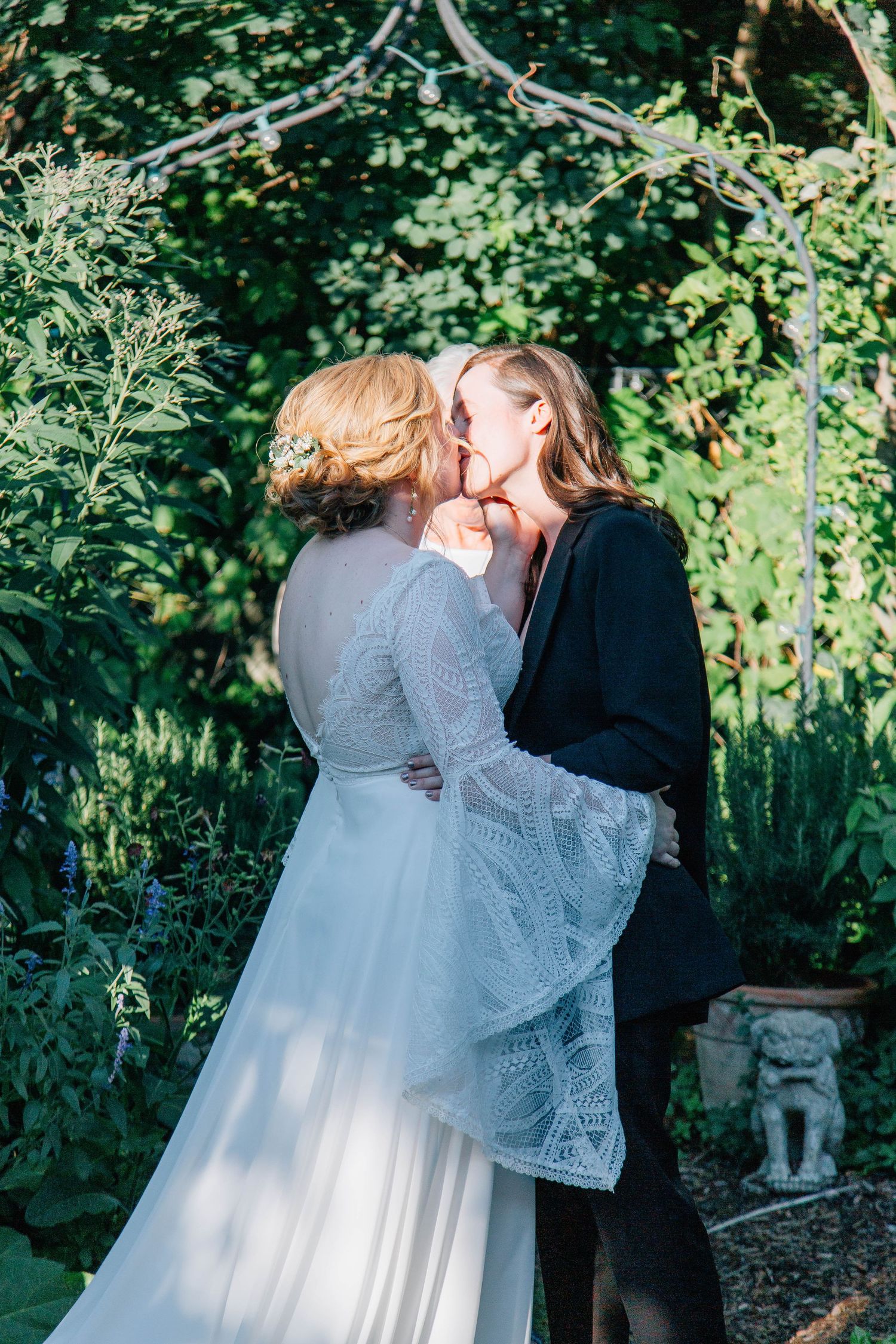 Two brides share first kiss surrounded by lush greenery during outdoor garden wedding ceremony.