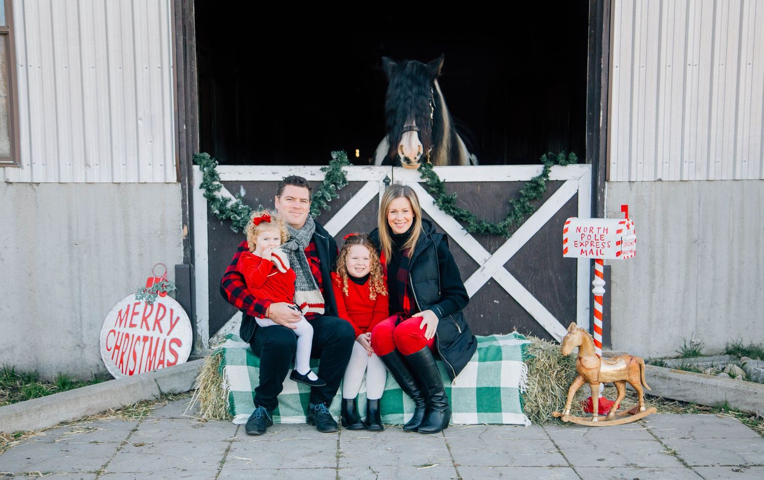 Family of four posing with horse at decorated barn for Christmas mini session.