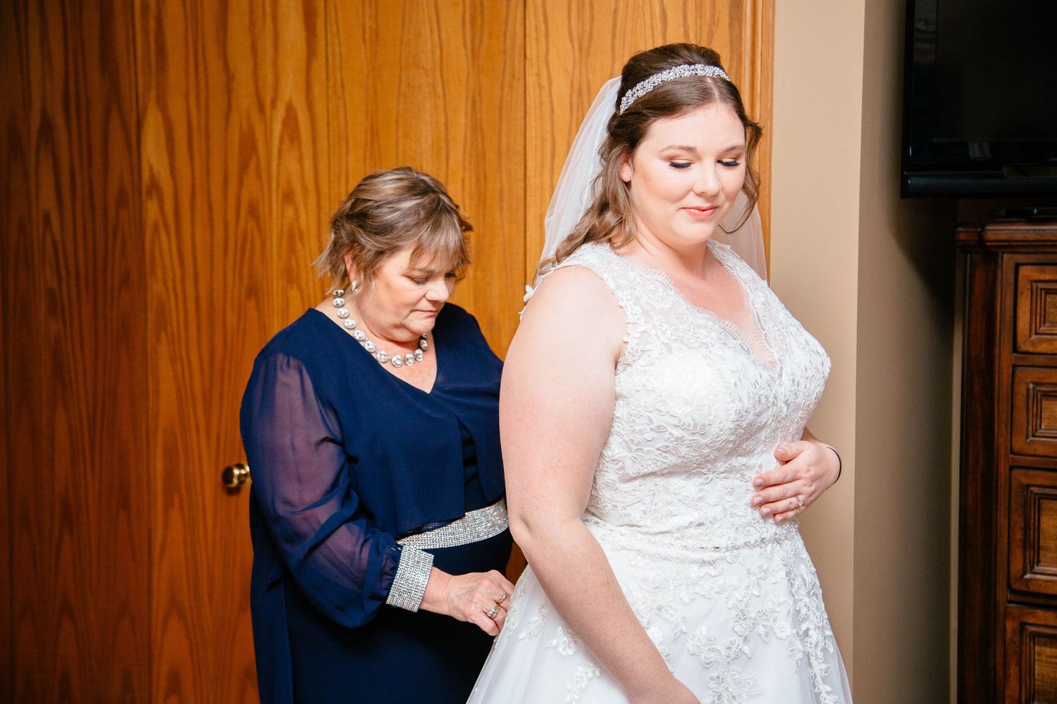 Mother in navy dress carefully adjusting brides white lace wedding gown belt during pre-wedding preparations indoors.