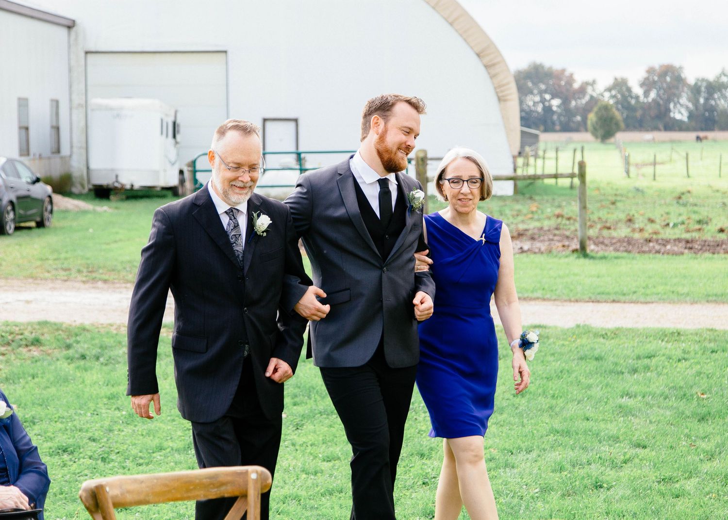 Groom in gray suit walks down the aisle escorted by his parents at an outdoor farm wedding ceremony.