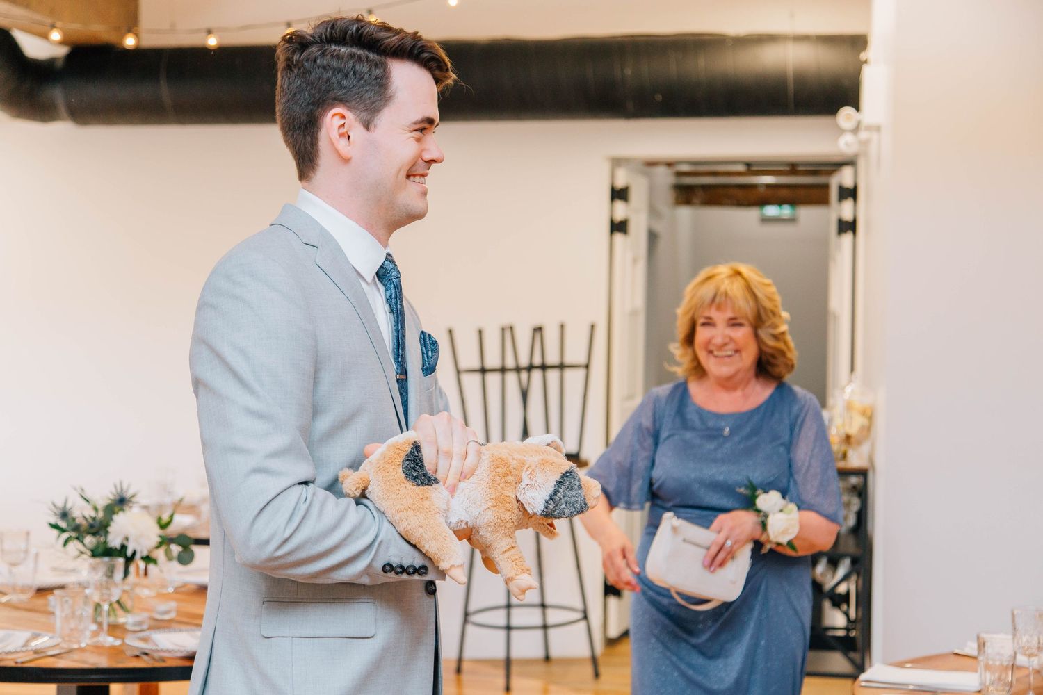 Smiling groom in gray suit holds stuffed animal gift from laughing woman in blue dress.