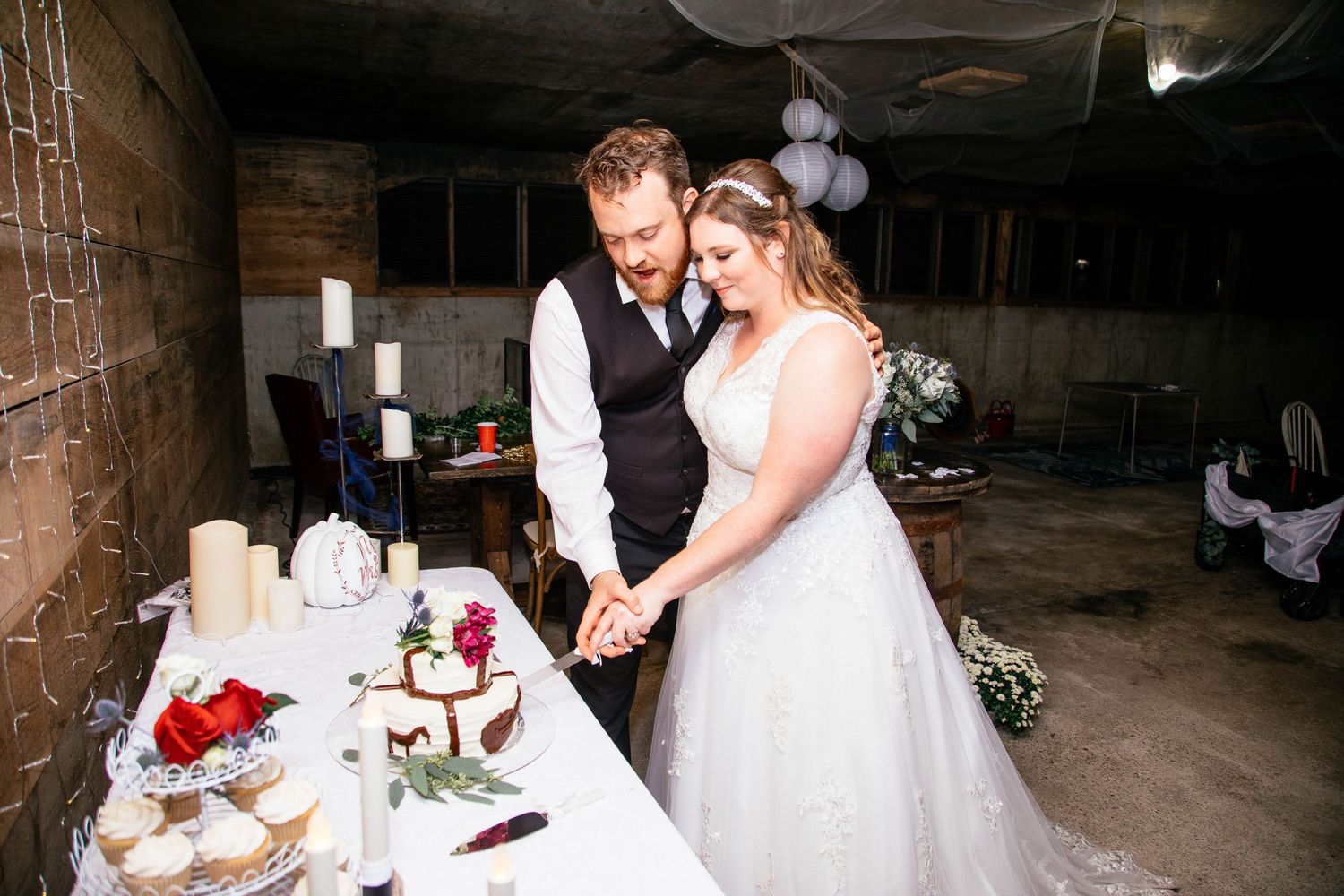 Bride and groom cutting their rustic wedding cake together at a candlelit reception table.