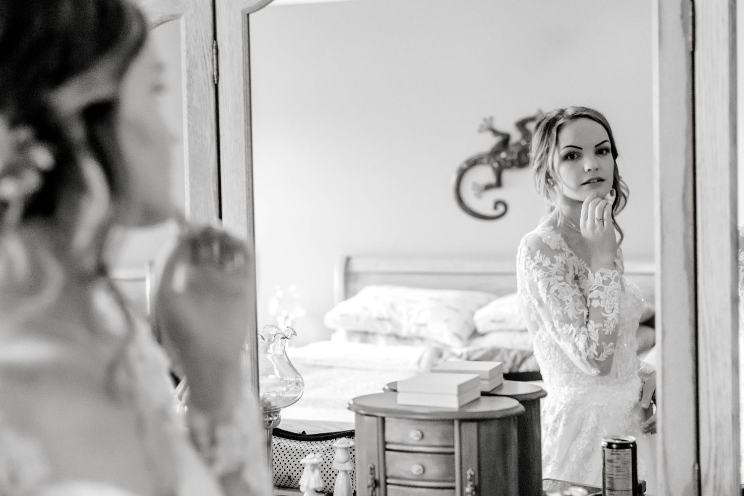 Black and white photo of bride in lace gown checking her makeup in a bedroom mirror.