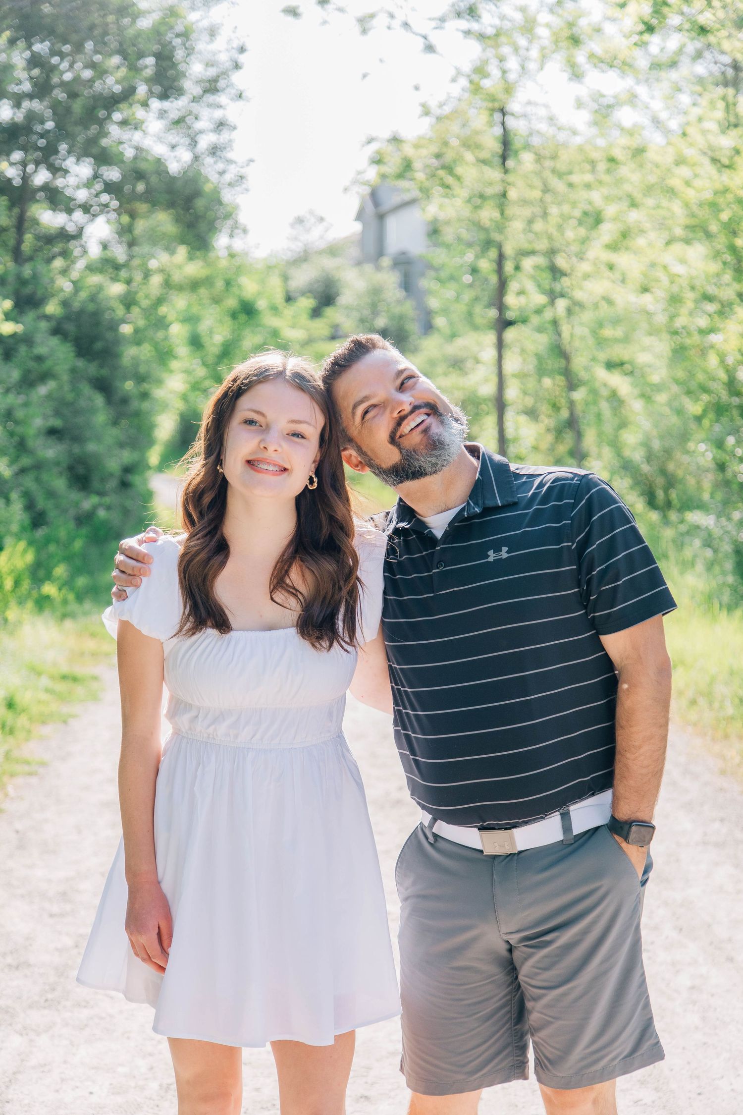 Smiling teenage girl and bearded father posing together on sunny outdoor gravel path.