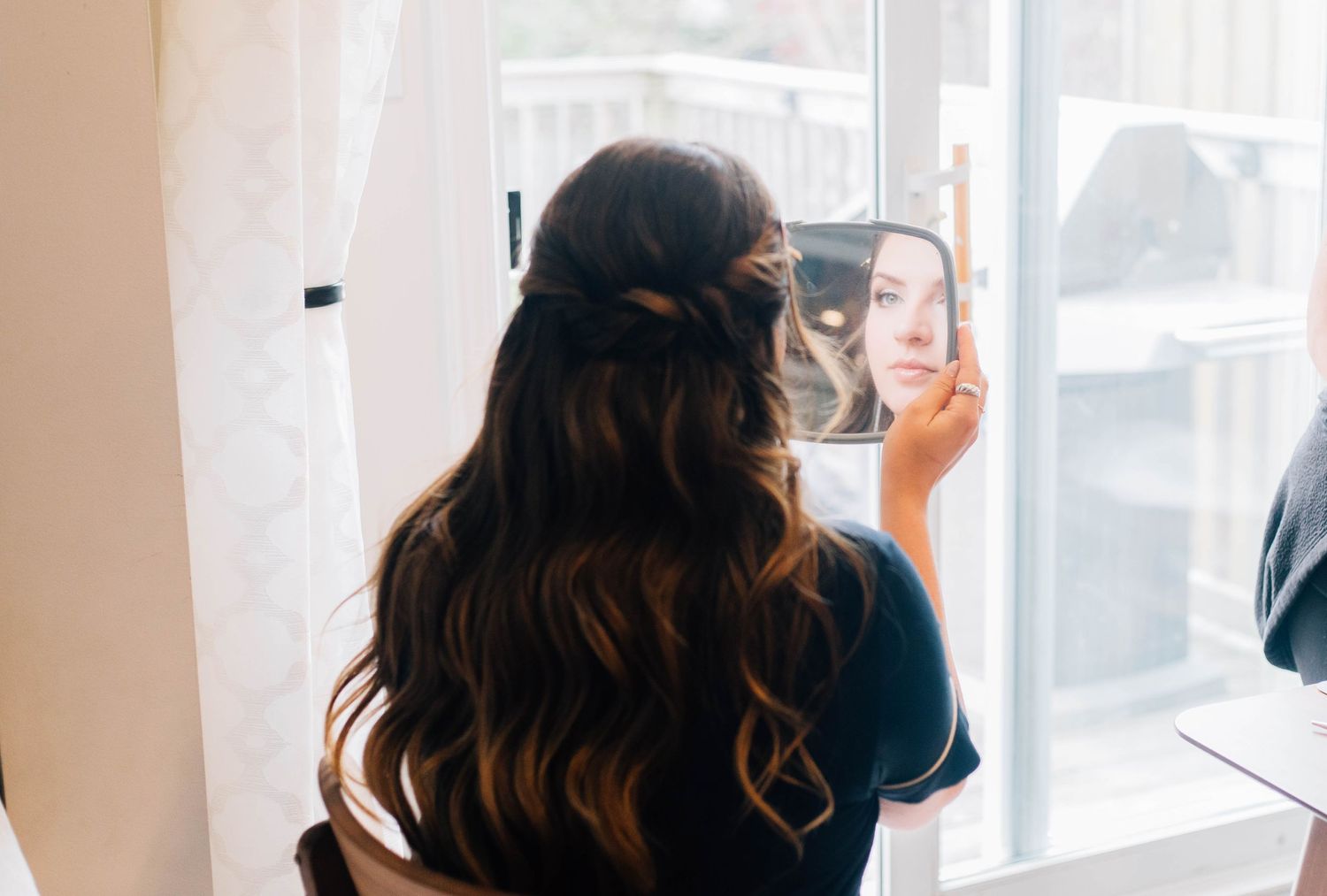 Bride with long dark hair checking her half-up hairstyle in handheld mirror by window.