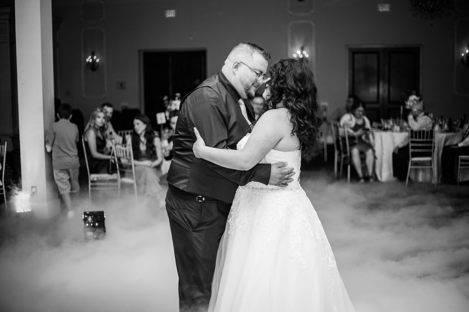 Black and white photo of bride and groom sharing first dance surrounded by fog at wedding reception.