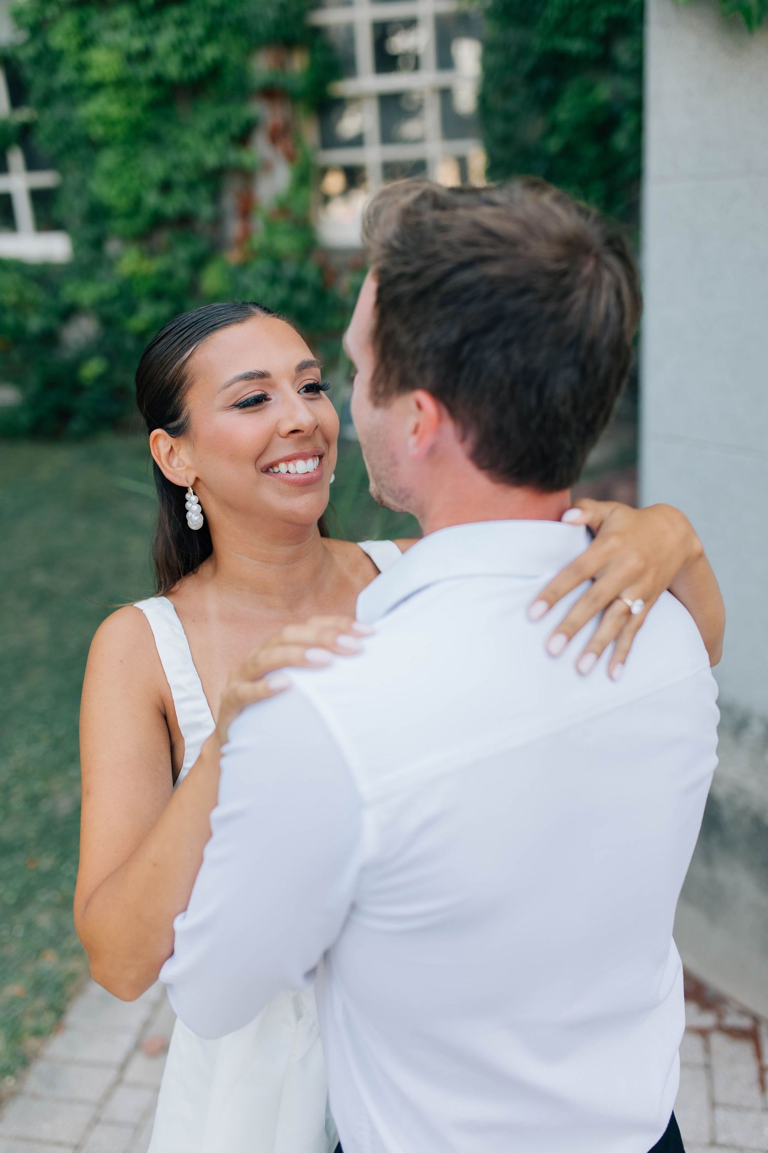 Bride smiles joyfully at groom while embracing outdoors near ivy-covered wall during engagement shoot.