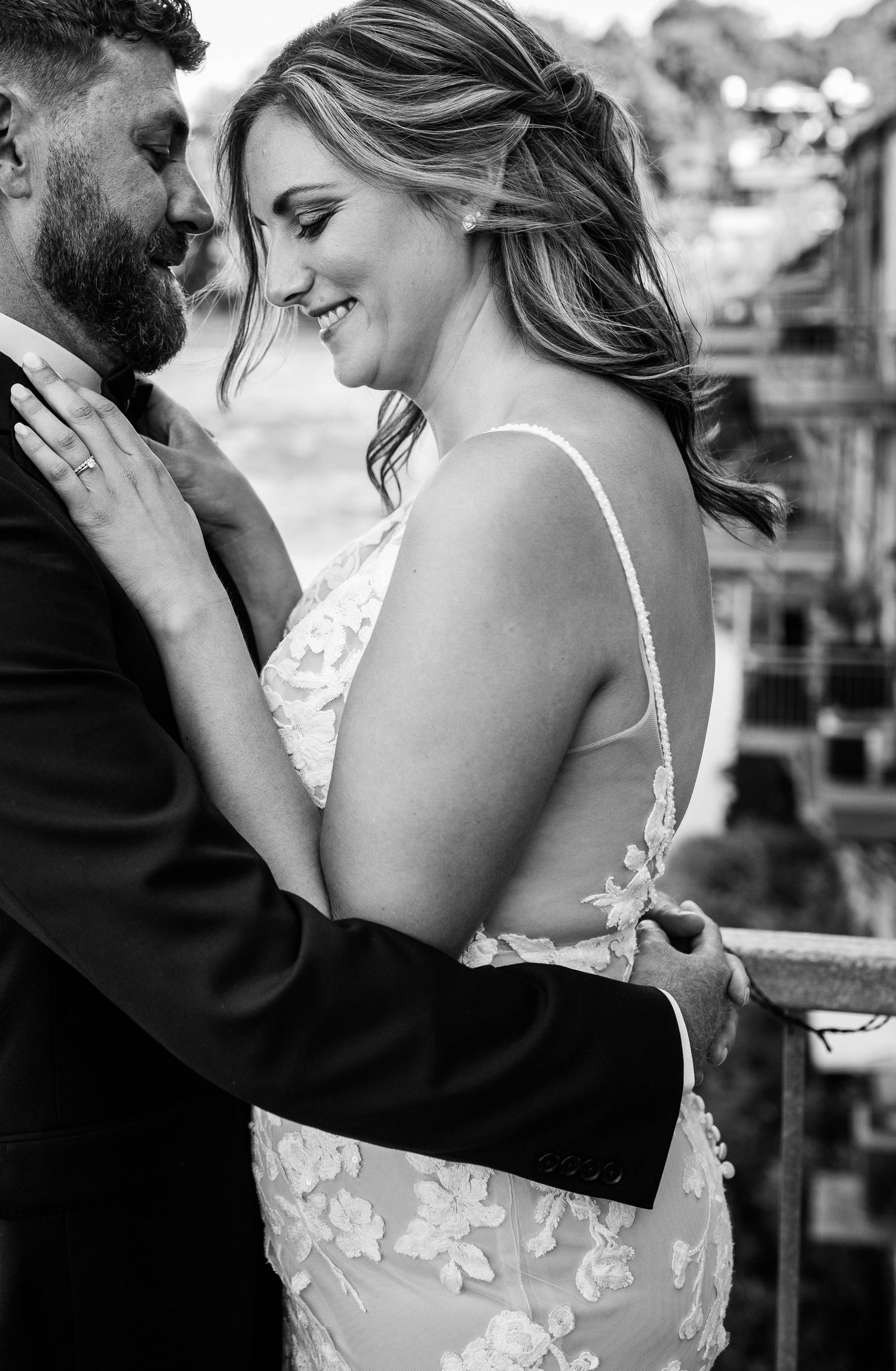 Black and white romantic portrait of smiling bride in lace gown and bearded groom on balcony.