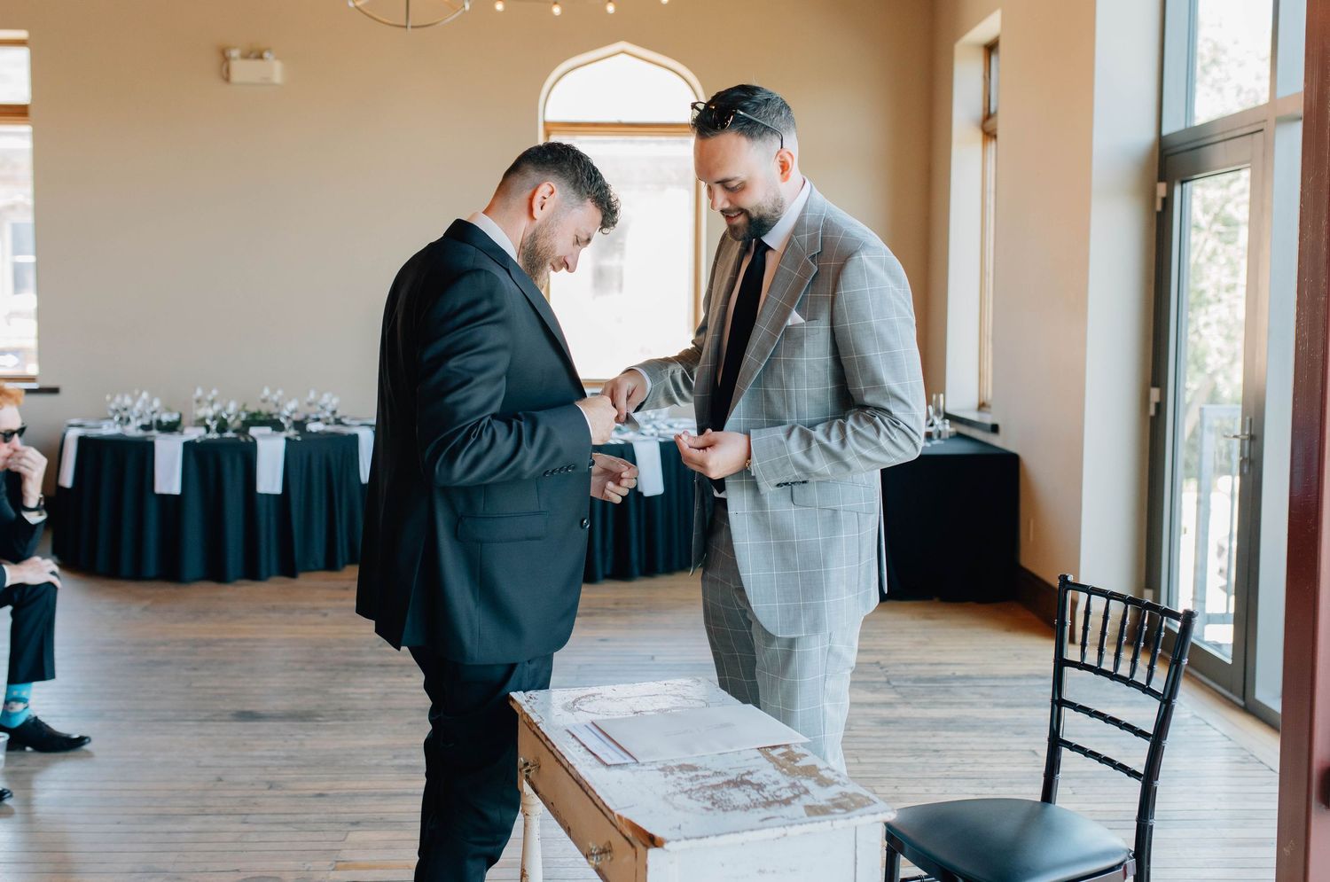 Two groomsmen in suits adjusting boutonnieres inside elegant wedding reception venue.