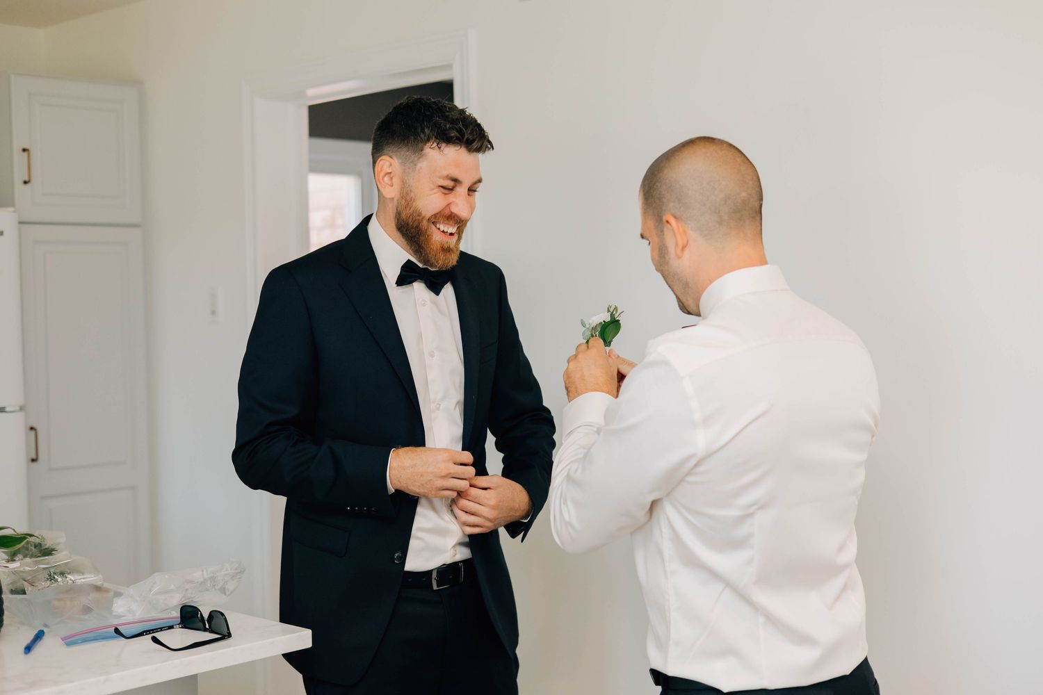 Groom in black tuxedo smiling as groomsman pins boutonniere on lapel indoors.