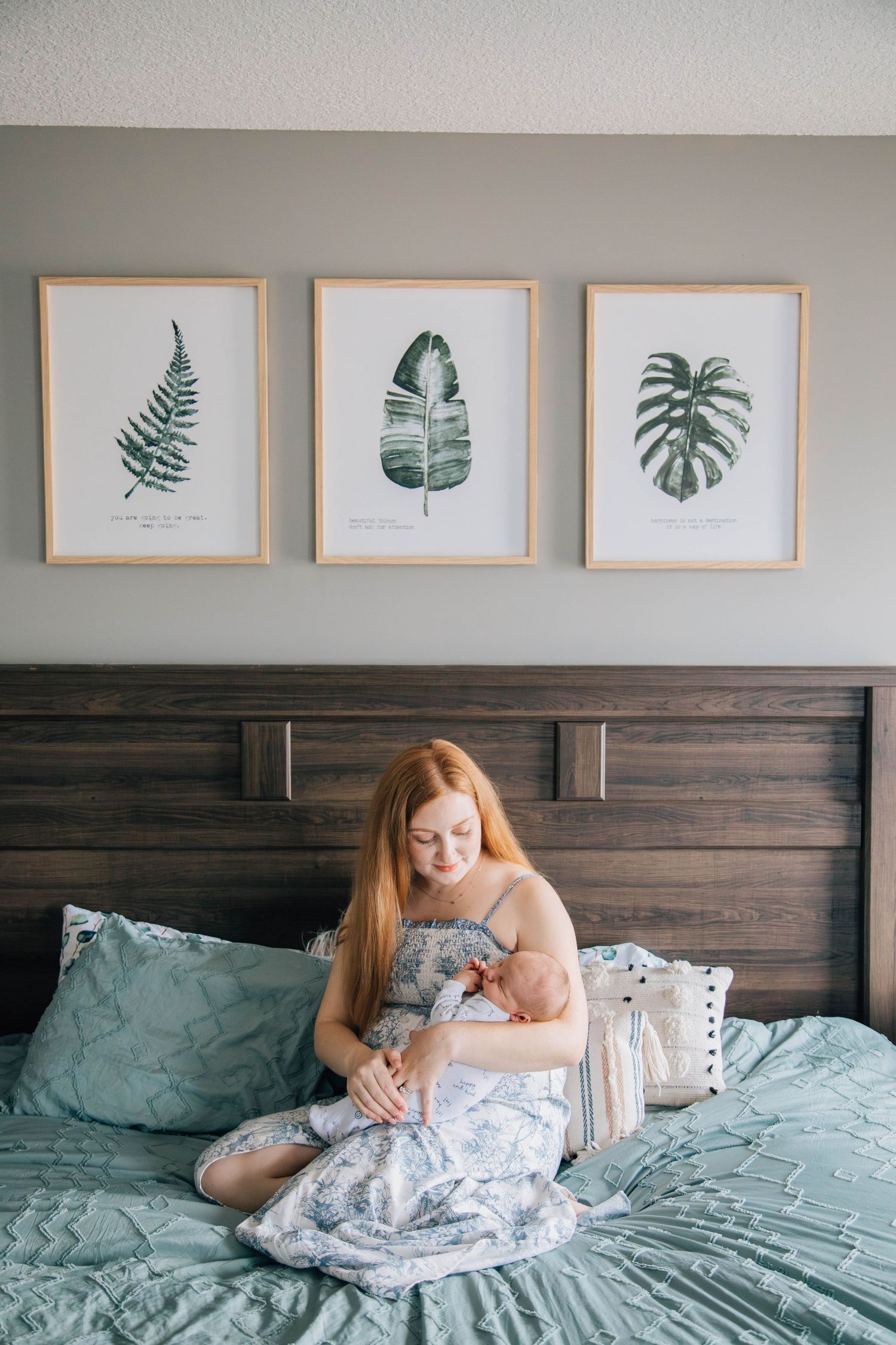 Redhead mother sitting on bed cradling newborn baby with botanical art prints on wall behind her.