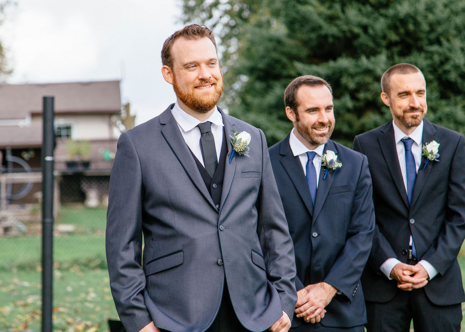 Groom in gray suit with boutonniere smiles at the altar flanked by groomsmen in navy suits at an outdoor wedding.