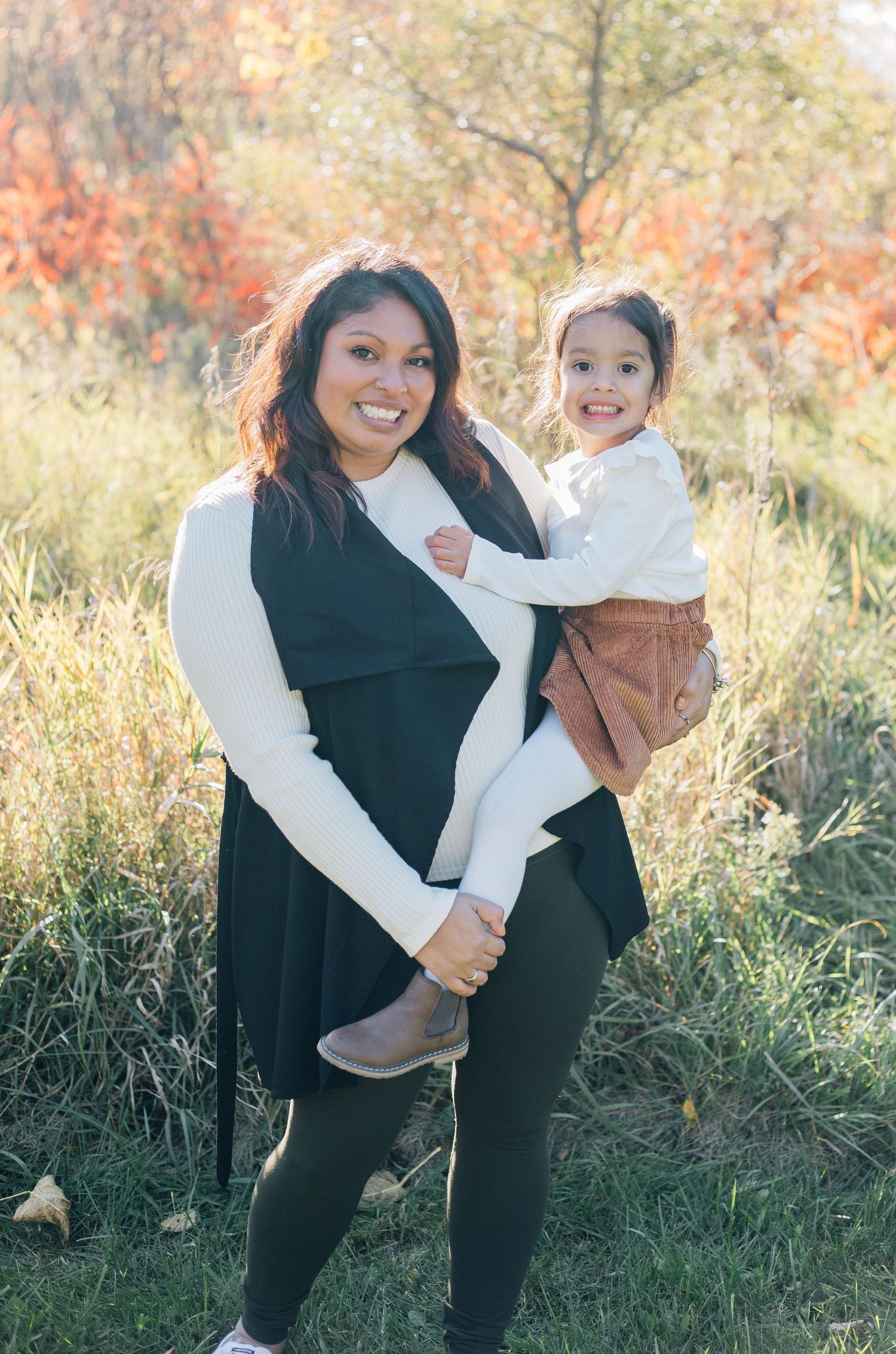Smiling mother holding young daughter outdoors in autumn foliage during family photo session.
