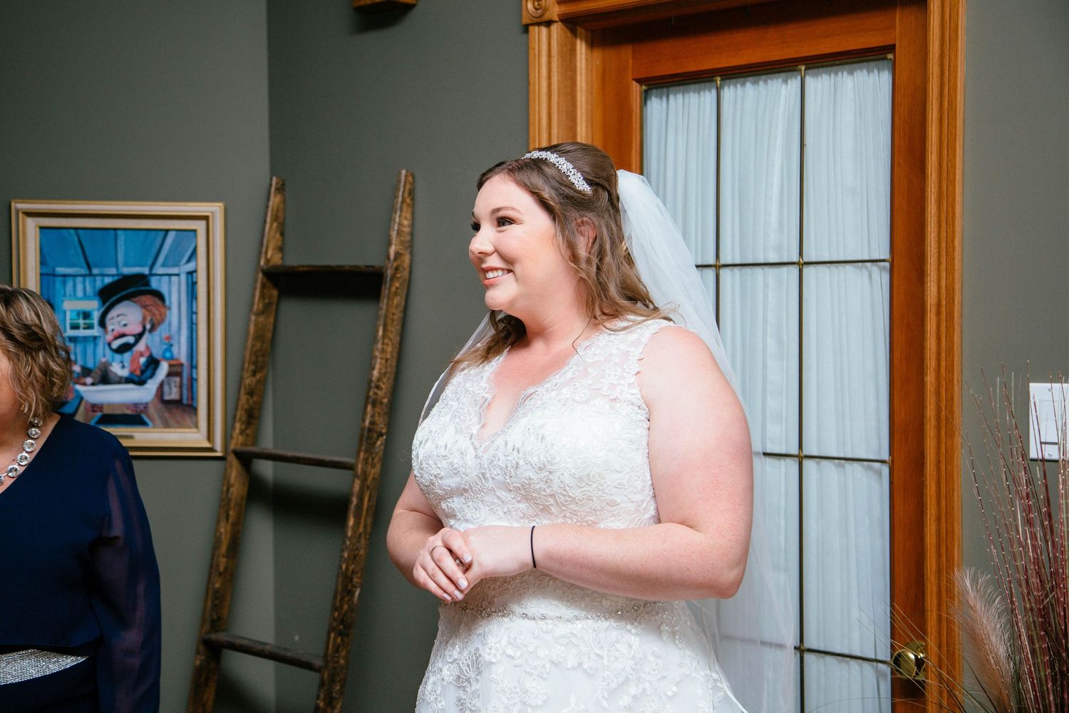 Bride in lace wedding gown and veil smiles warmly while standing in a rustic venue doorway.