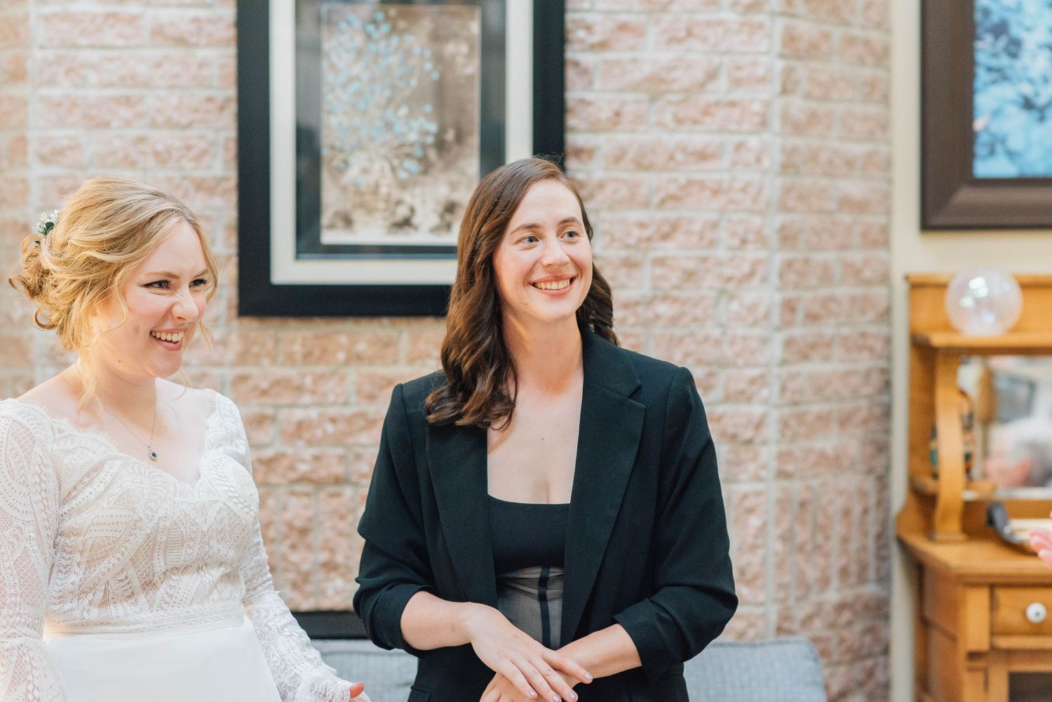 A bride in white and a woman in a black blazer smiling together at a wedding.