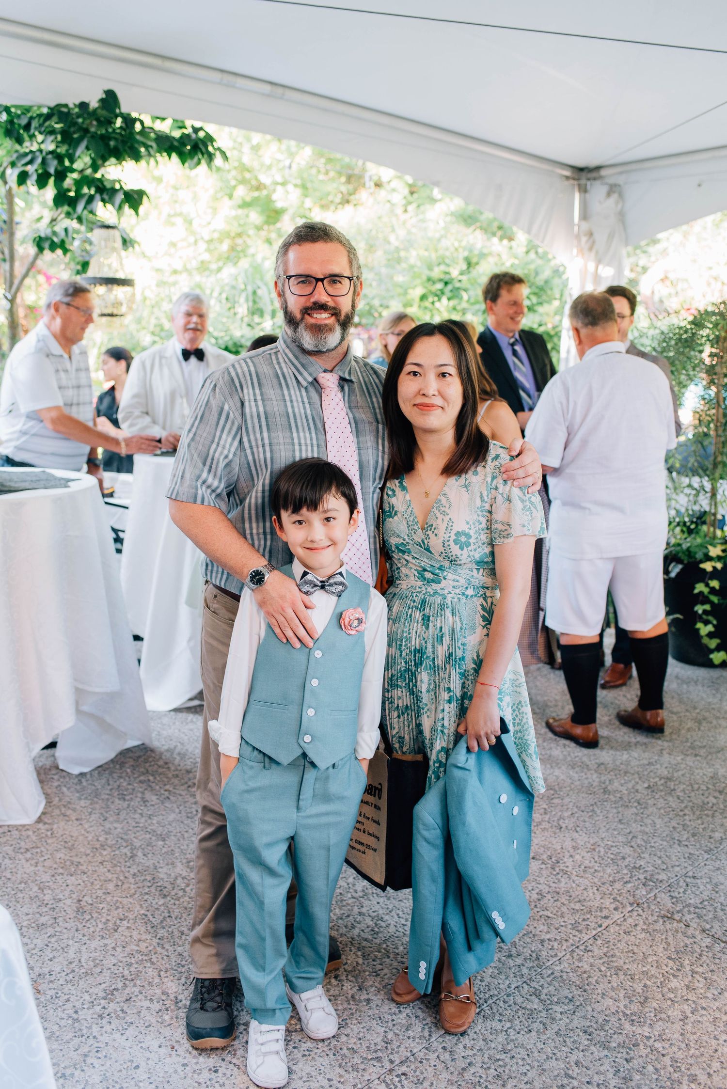 Family of three poses together under tent at outdoor wedding reception in summer garden.