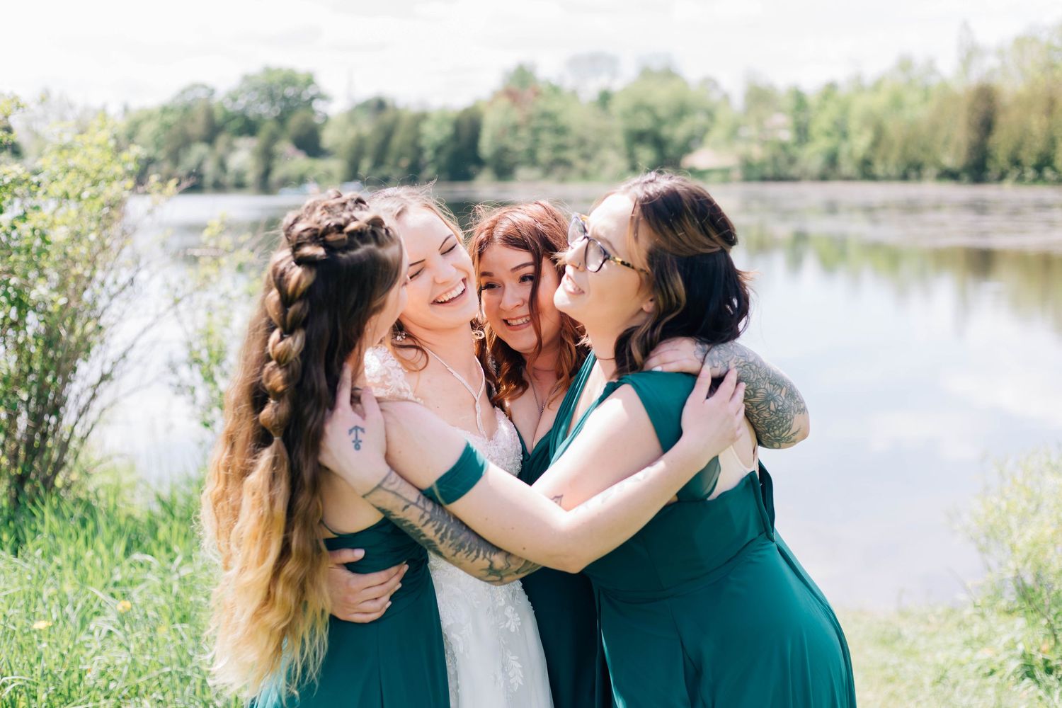 Bride and three bridesmaids in emerald dresses sharing a joyful group hug outdoors.