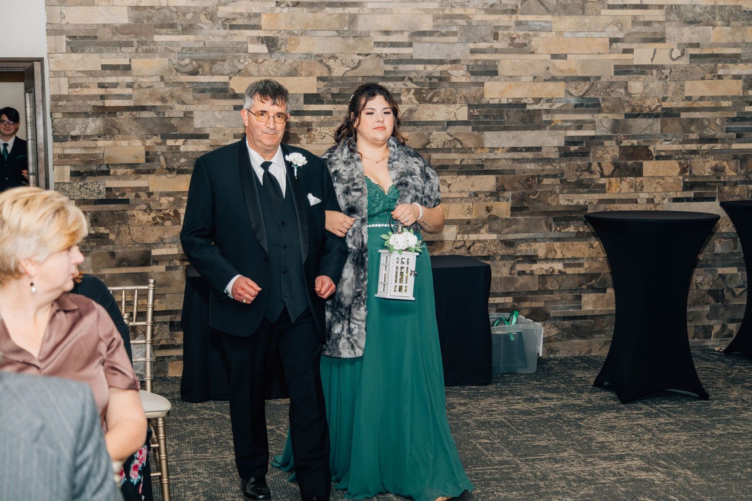 Bridesmaid in green gown carrying lantern walks with groomsman past seated guests at winter wedding ceremony.