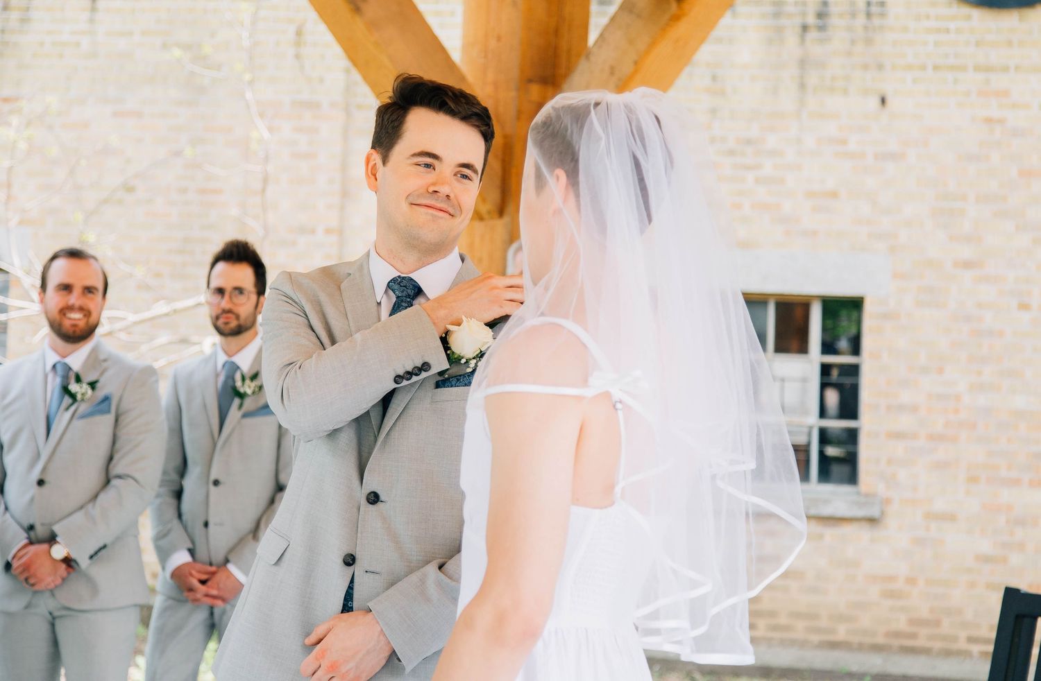 Emotional groom in gray suit lifts bride's veil during outdoor wedding ceremony.