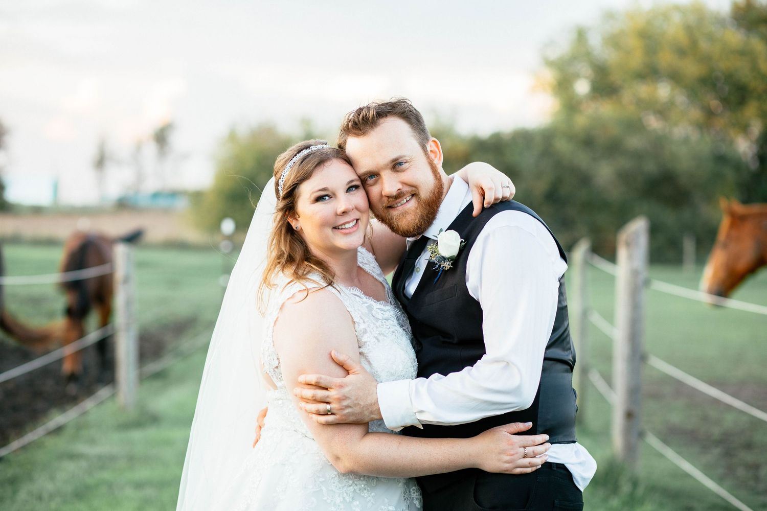 Smiling bride and groom embracing outdoors with horses grazing in the background.