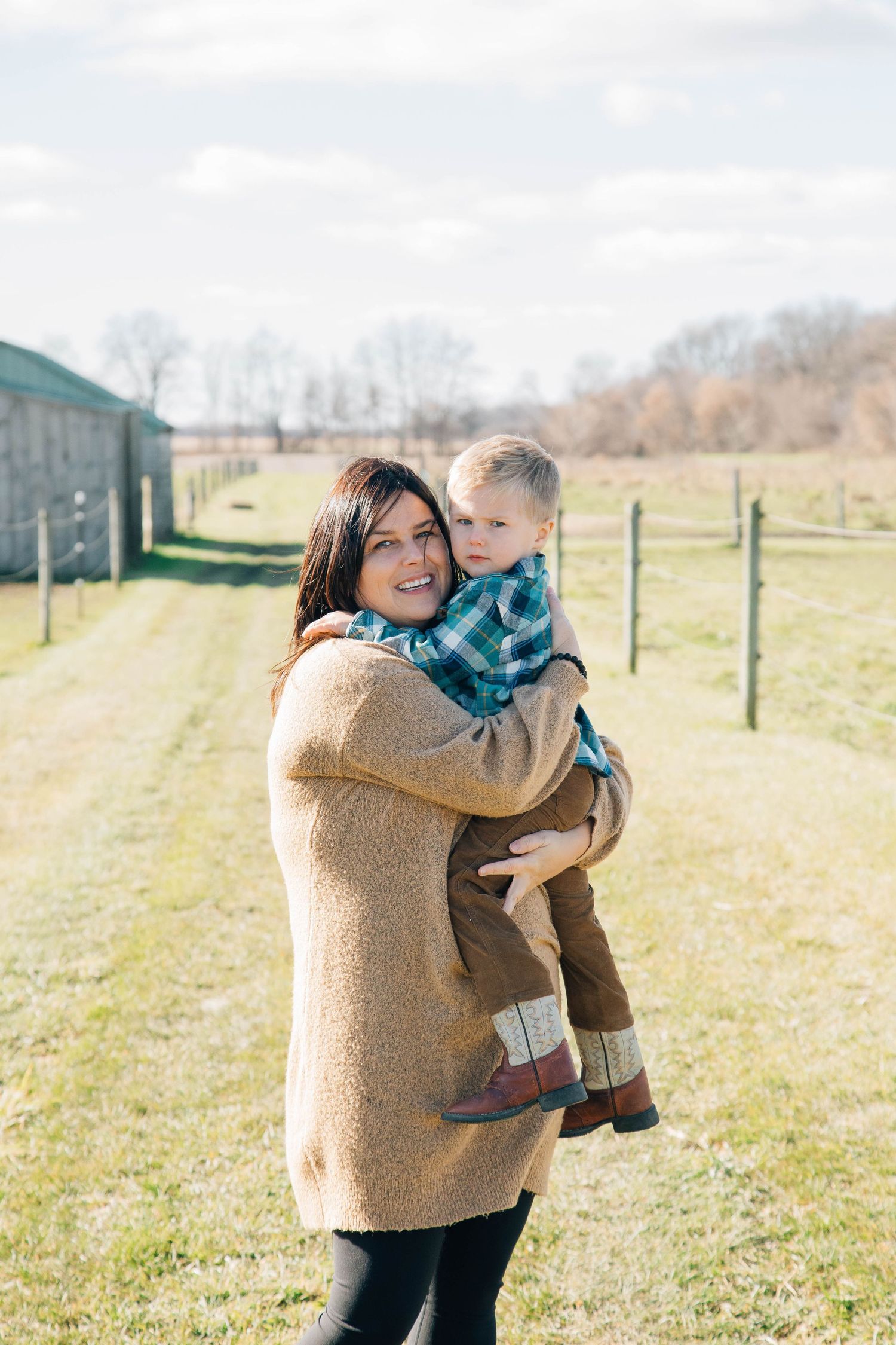 Smiling mother holding young toddler son in plaid shirt on sunny farm field.