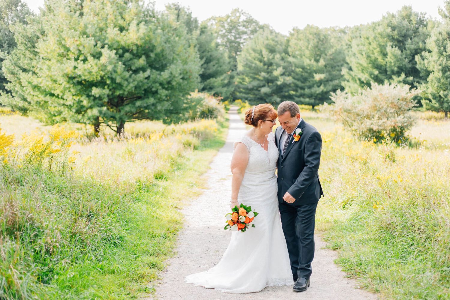 Bride and groom walking on a path surrounded by lush green trees.