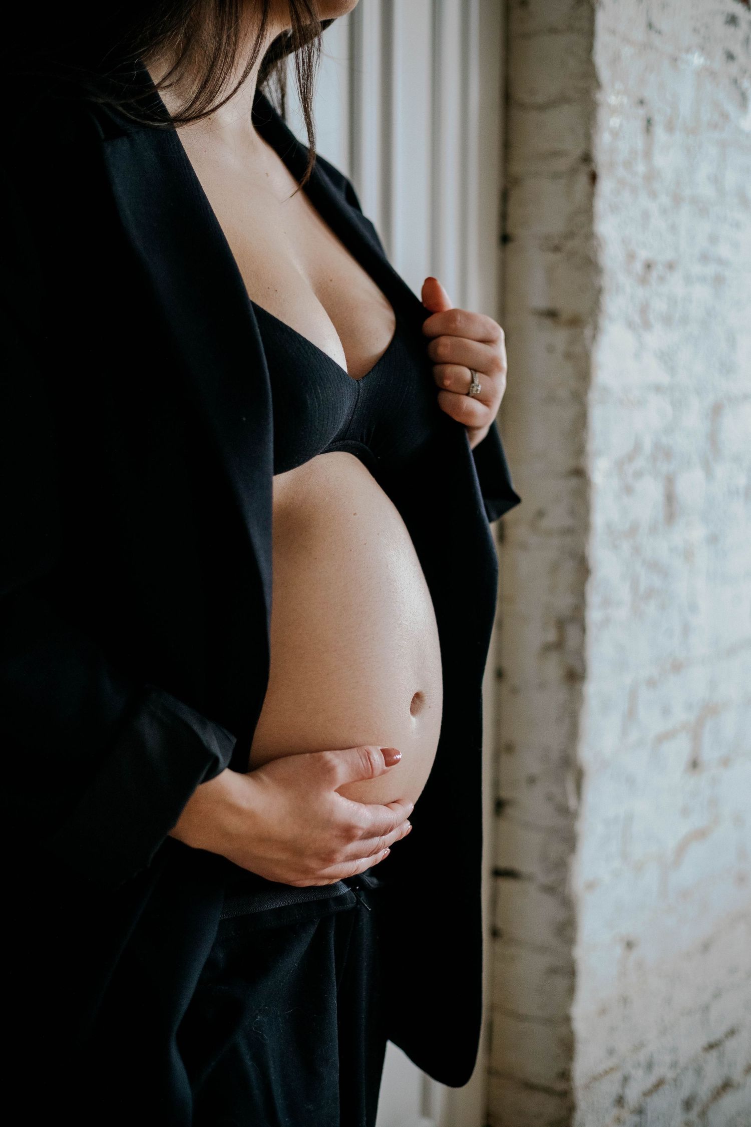 Close-up of a pregnant woman in a black blazer cradling her bare baby bump with both hands.