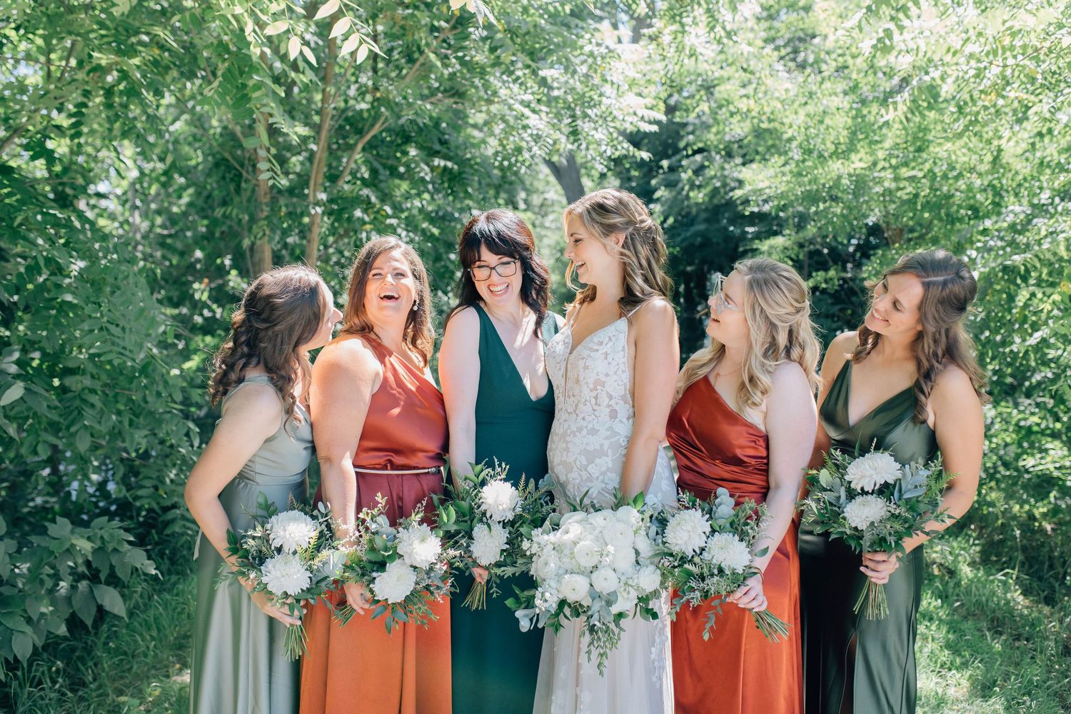Bride and bridesmaids in earth-tone dresses laughing together outdoors on a trail in Paris Ontario.