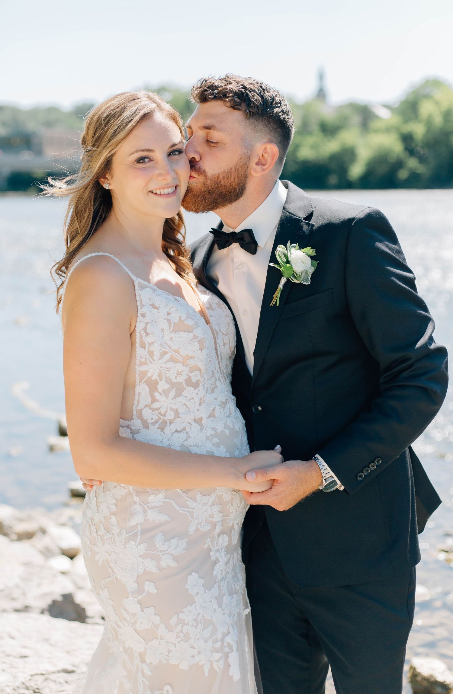 Groom in black tuxedo kisses bride in lace gown on the cheek near a sunlit waterfront in Paris Ontario.