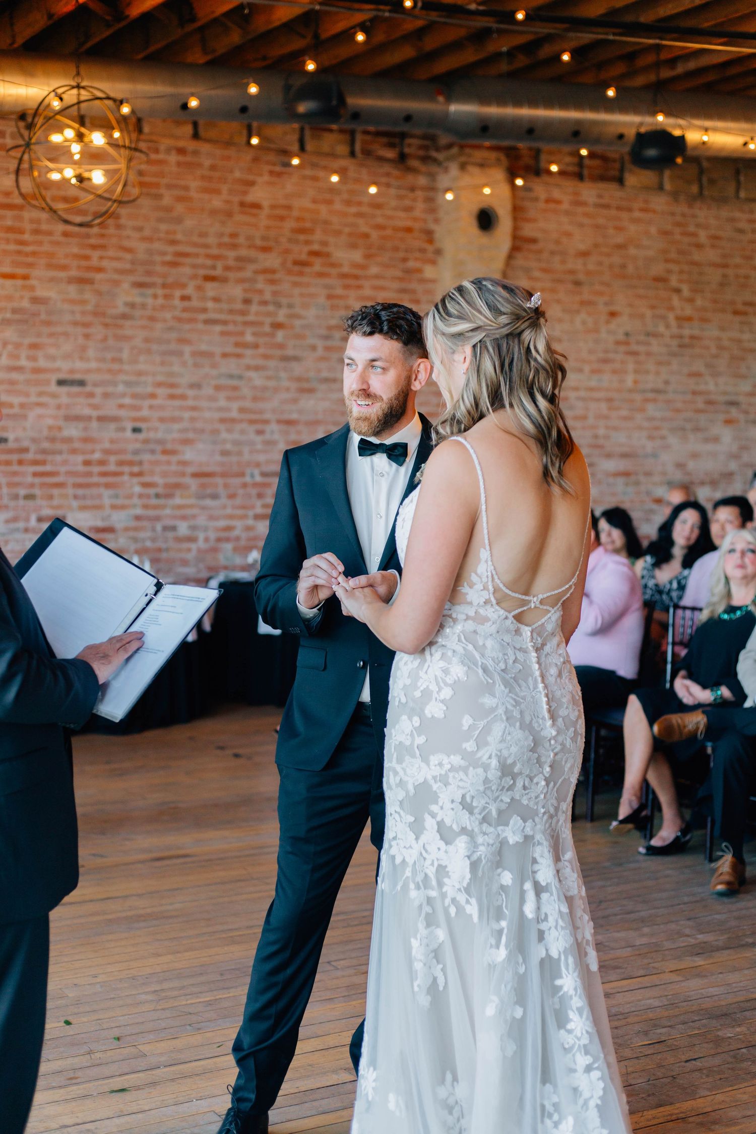 Groom in black tuxedo placing ring on brides finger during wedding ceremony in rustic brick venue.