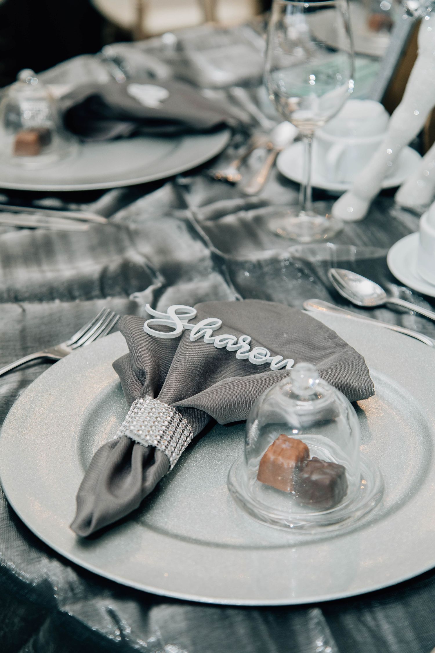 Elegant winter wedding place setting with gray napkin, rhinestone ring, laser-cut name tag, and chocolate under glass dome.