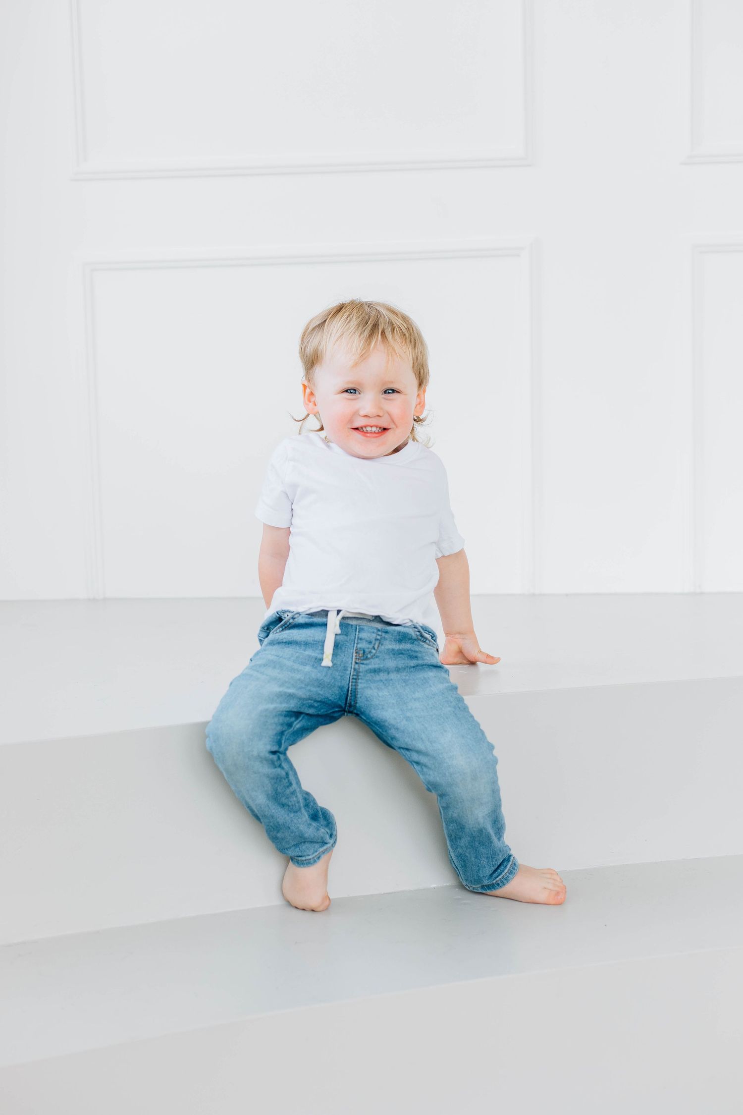 Smiling toddler boy in white t-shirt and jeans sitting on white surface in bright studio.