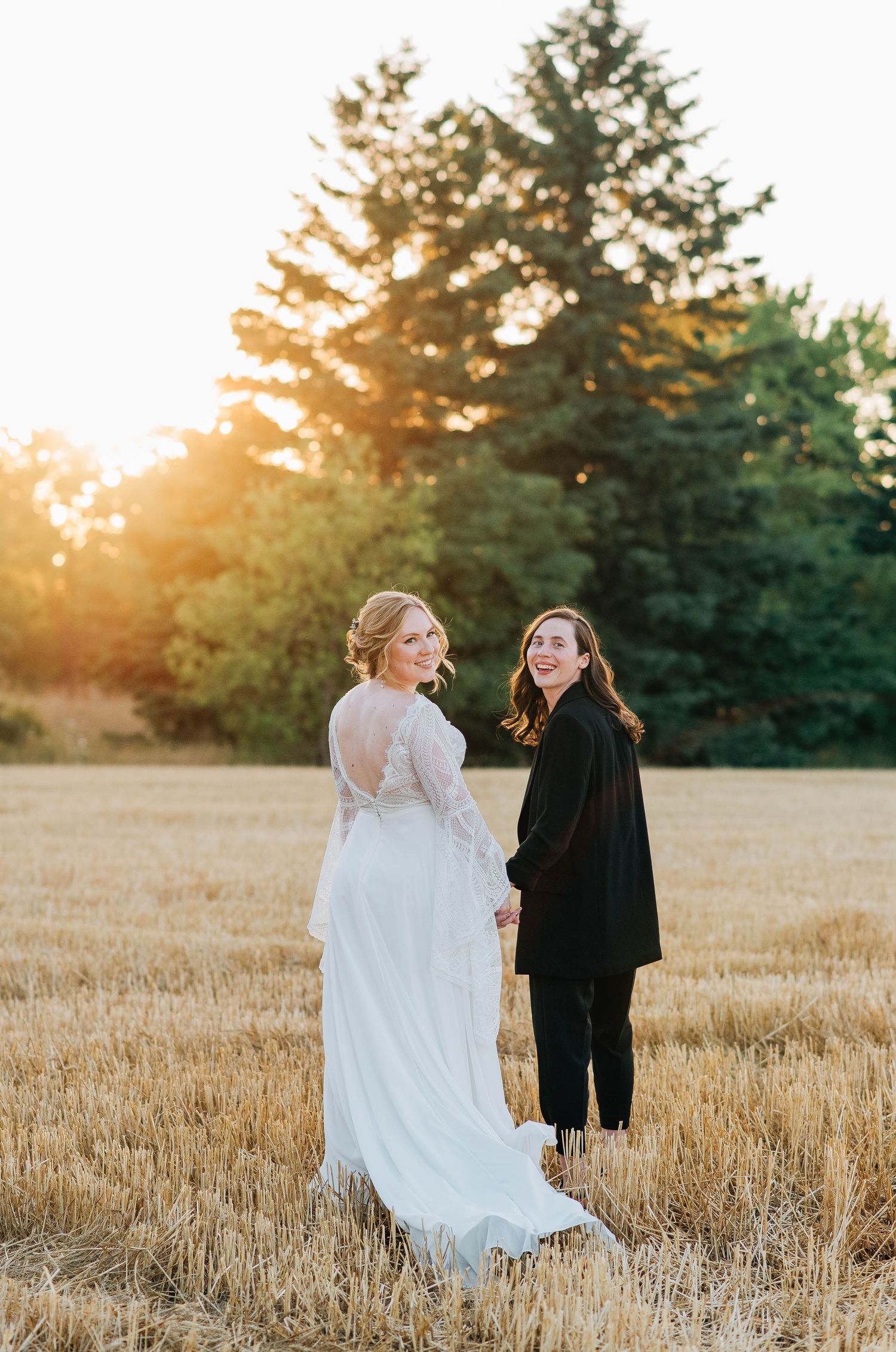 Two brides holding hands in golden wheat field at sunset, one in white lace gown with train, one in black.